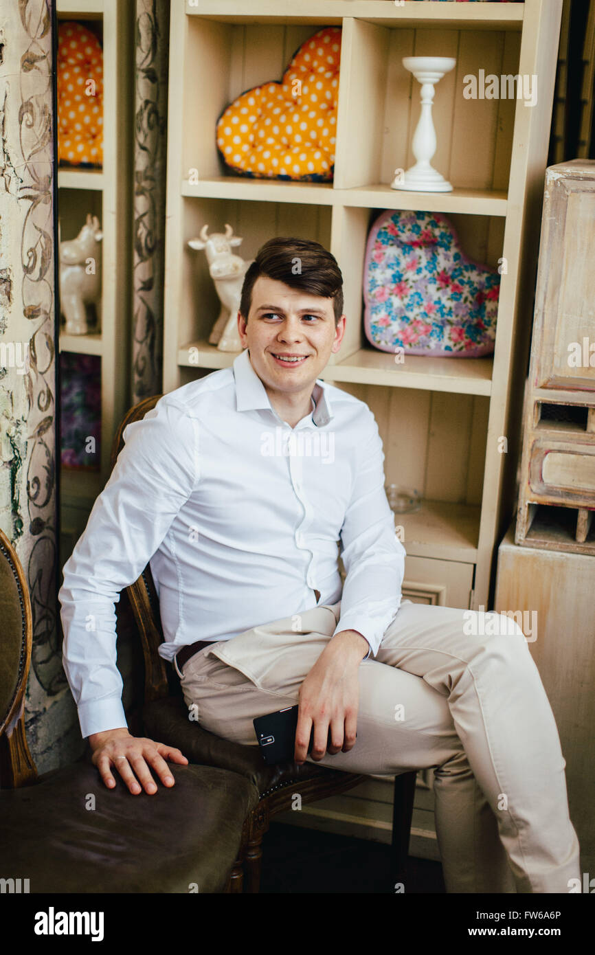 Ritratto emotivo di sorridere bello hipster in bianco shirt casual seduto su una sedia vintage con uno smartphone in stile retrò mi Foto Stock
