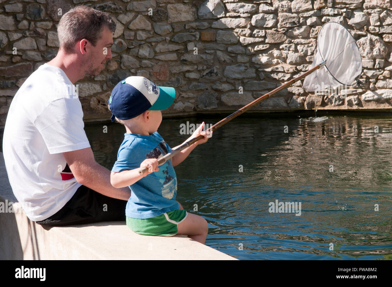 Padre e figlio seduta sul bordo di una piscina a dondolare i piedi nell'acqua giocando con una rete da pesca Foto Stock