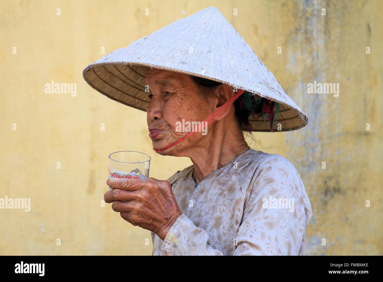 Ritratto di signora rugosa con cappello conico avente un drink, Hoi An, Vietnam Asia Foto Stock
