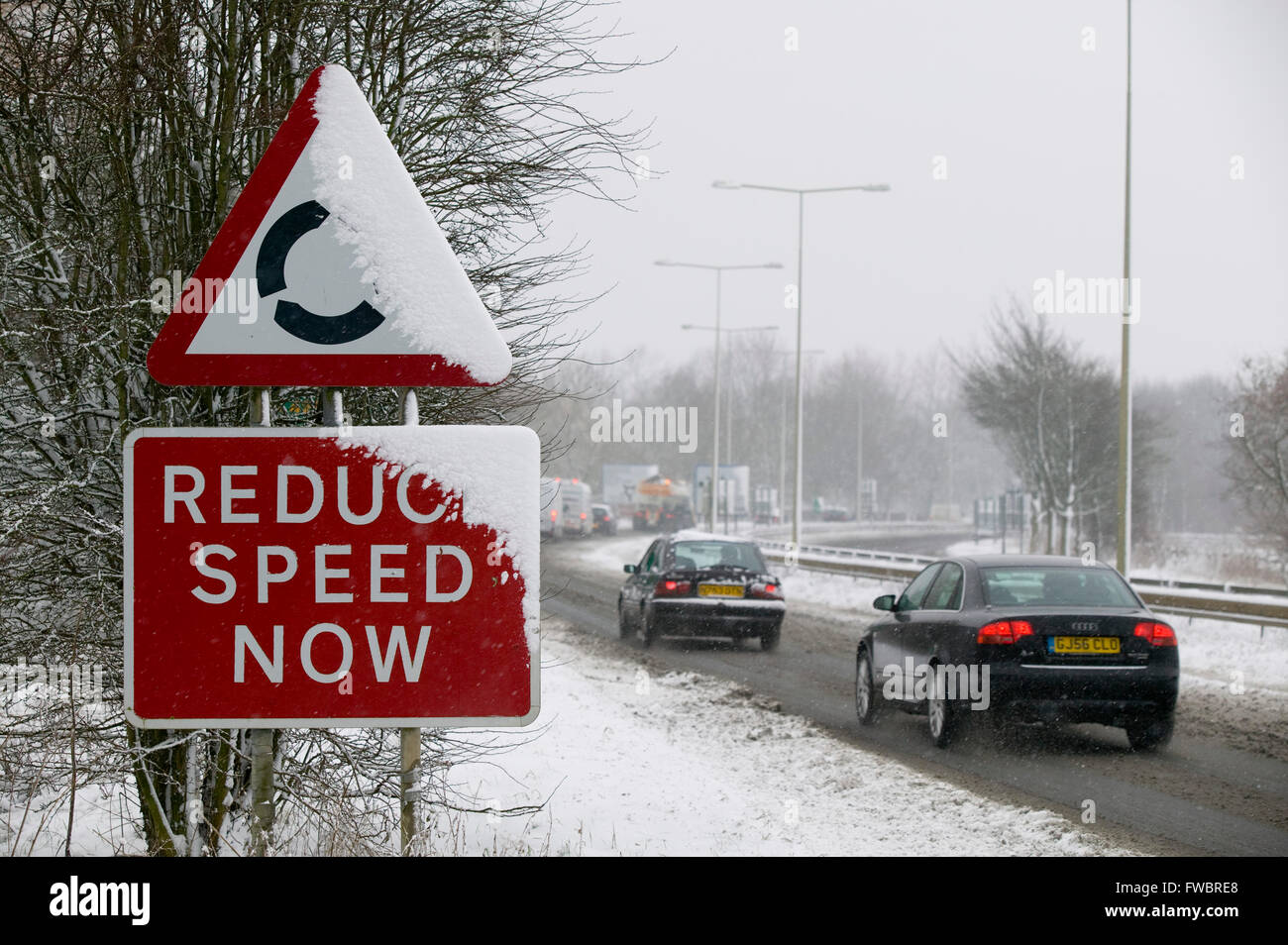 Un traffico stradale segno ricoperta di neve di avvertimento di un su e venire rotonda e raccontare ai conducenti di ridurre la loro velocità. Le istruzioni di essere ancor più pertinenti in questa caduta di neve durante un inverno britannico dove le strade sono ghiacciate, scivoloso e molto pericolosa. Foto Stock