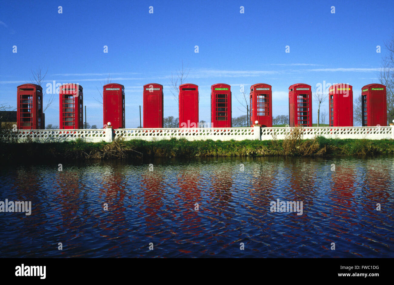 Raccolta di scatole telefoniche rosse allineate e adagiato sul bordo di un laghetto che riflette il colore e la forma dell'acqua. Foto Stock