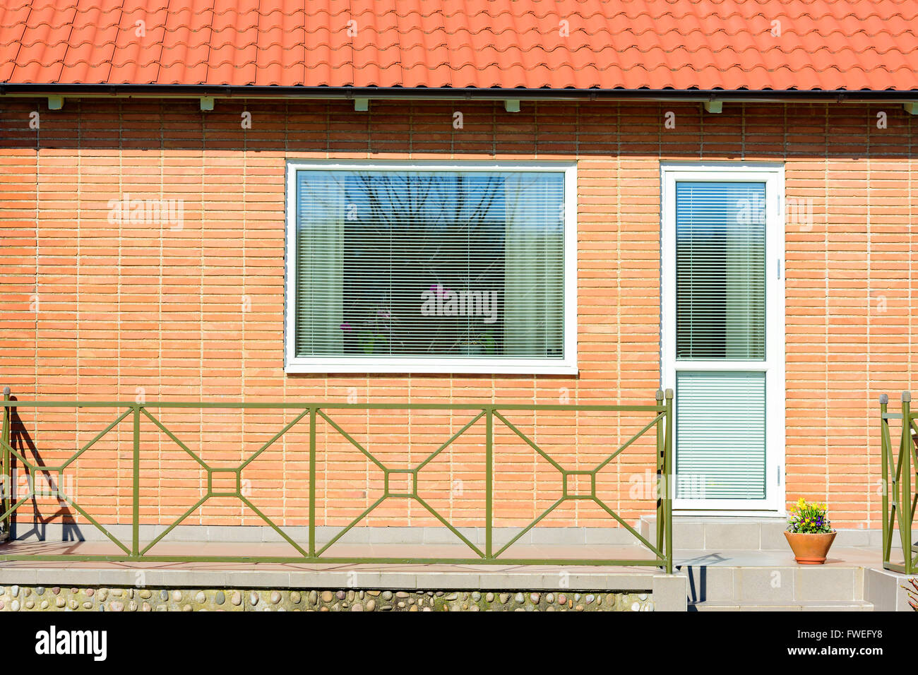 Dettaglio di una facciata di mattoni rossi con una finestra e una porta di patio. Fiori e tenda in finestra, blinders verso il basso. Ringhiera metallica in fron Foto Stock