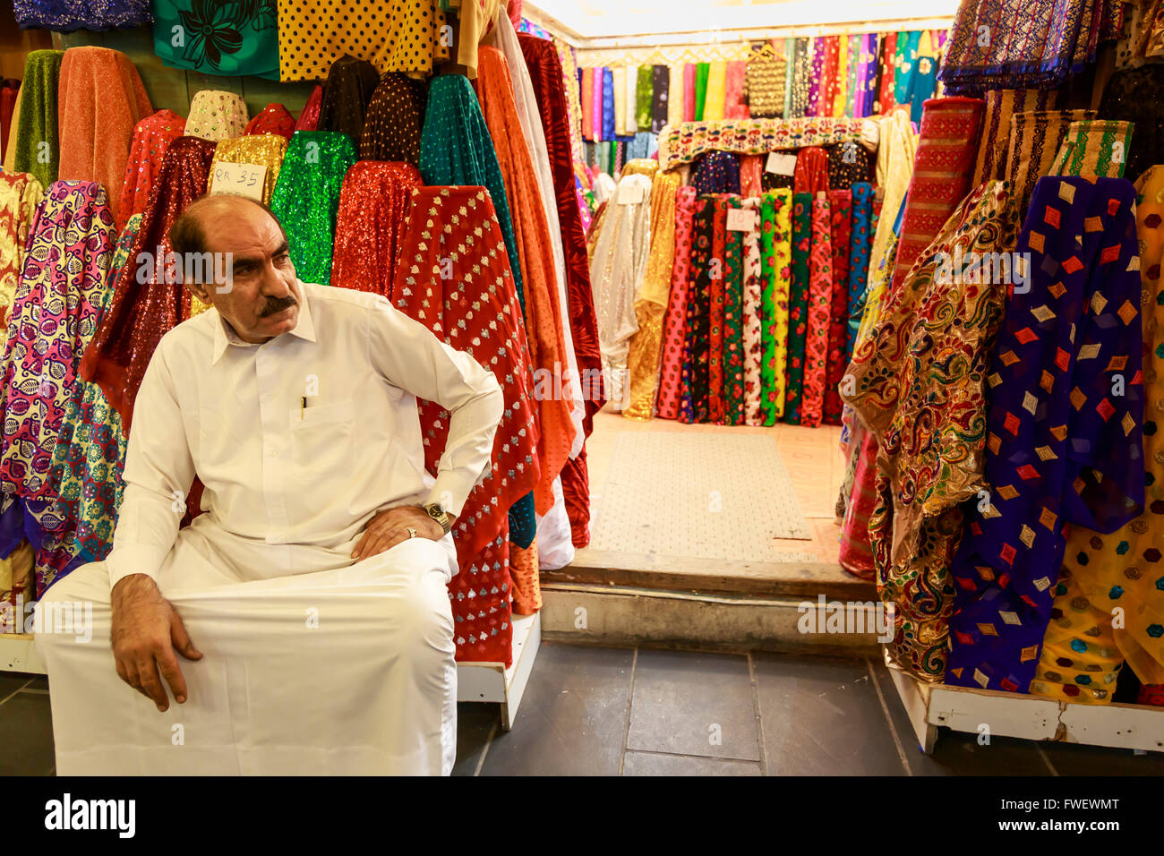 Negoziante seduto al di fuori del suo negozio circondato da materiale colorato, Souq Waqif, Doha, Qatar, Medio Oriente Foto Stock