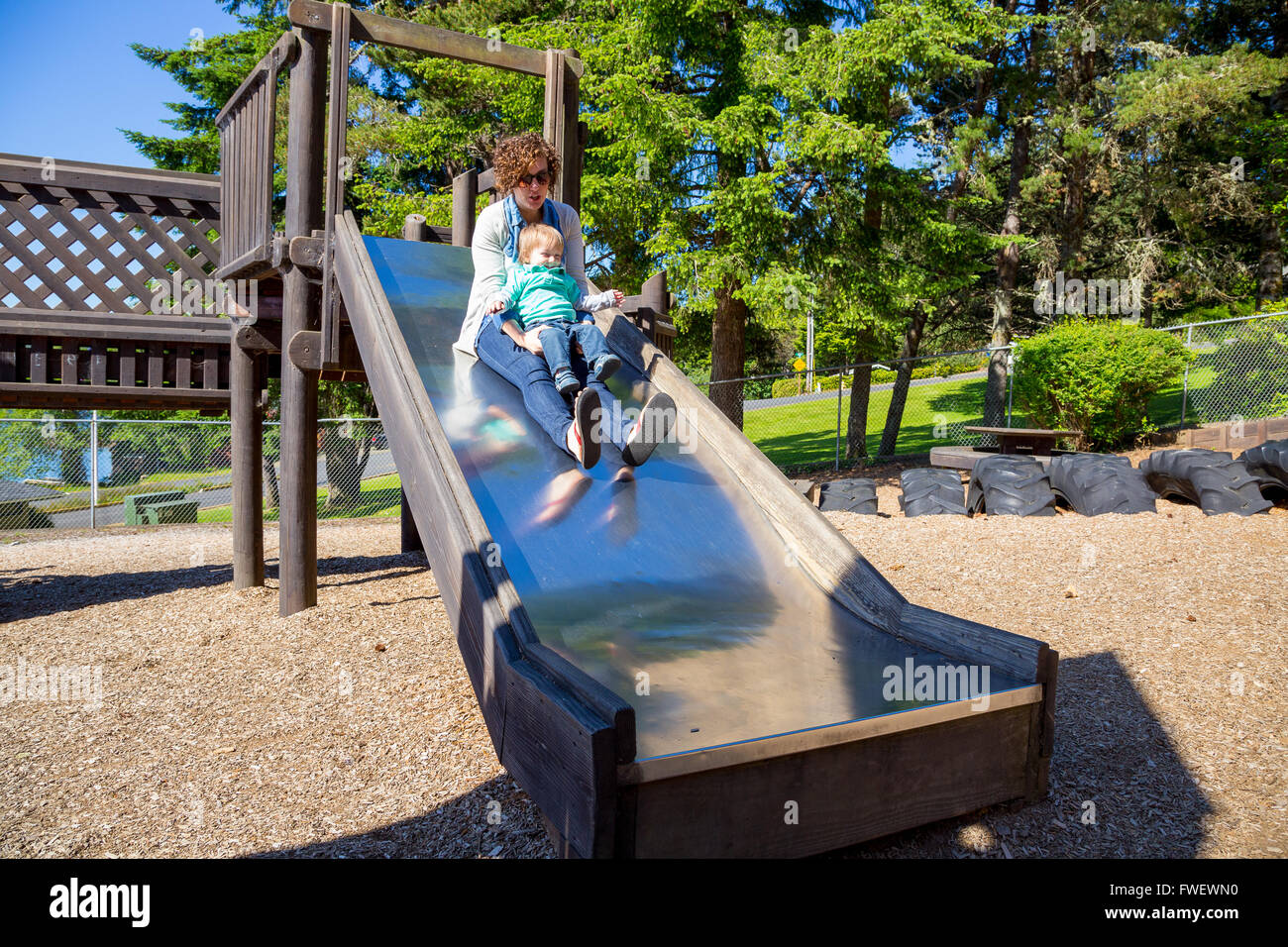 Una madre e suo figlio far scorrere in basso la slitta in metallo in corrispondenza di un parco giochi all'aperto a un parco. Foto Stock