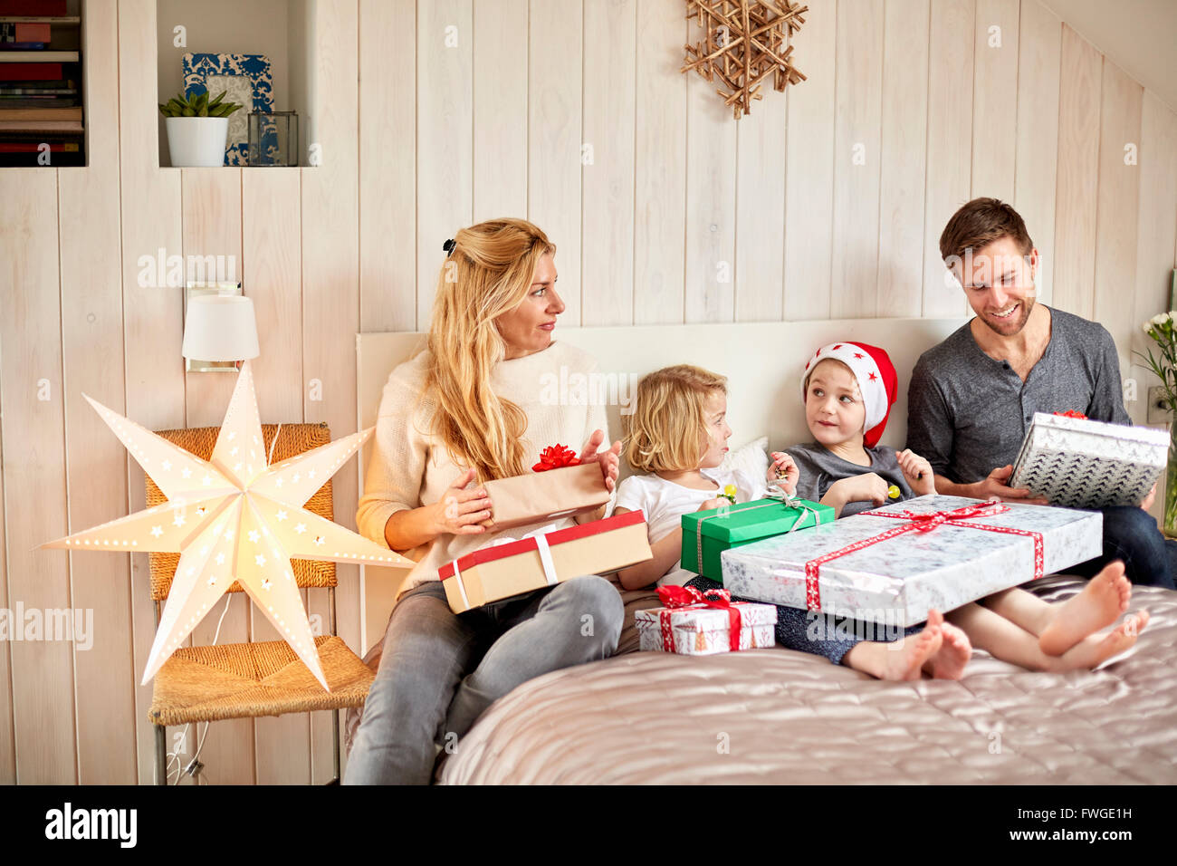 Una famiglia di due adulti e due bambini seduti nel letto la mattina di Natale apertura presenta insieme. Foto Stock