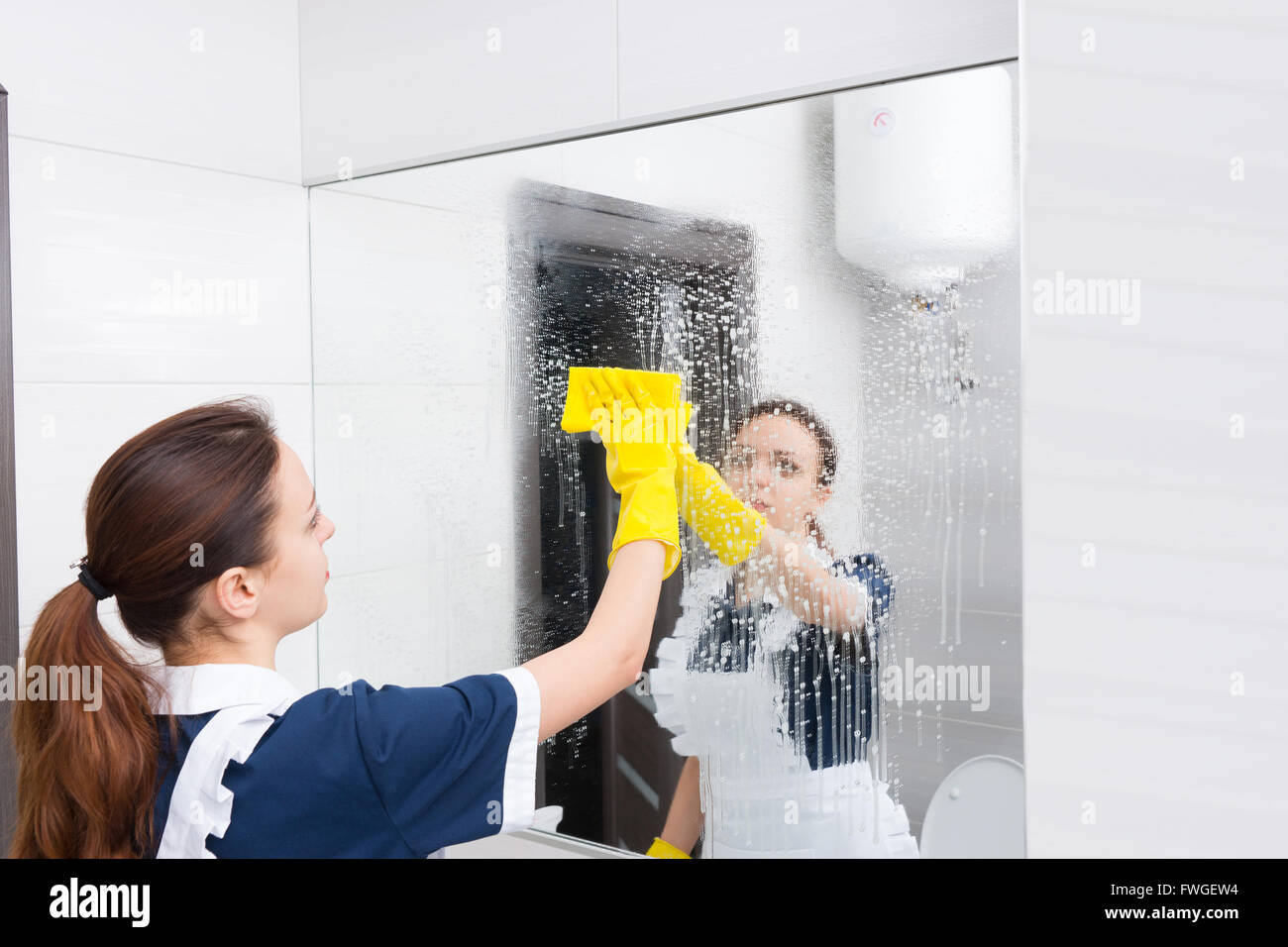 Governante di pulizia o la pulizia di una grande parete a specchio in un bagno bianco di strofinamento con un panno dopo la spruzzatura con il detersivo Foto Stock