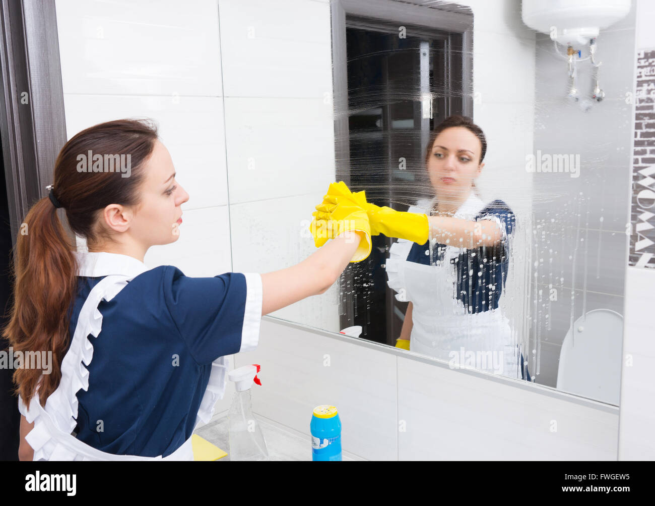 Hotel sporco lo specchio del bagno da ripulire con giallo rag dalla cameriera in blu e bianco uniforme e giallo guanti di gomma Foto Stock