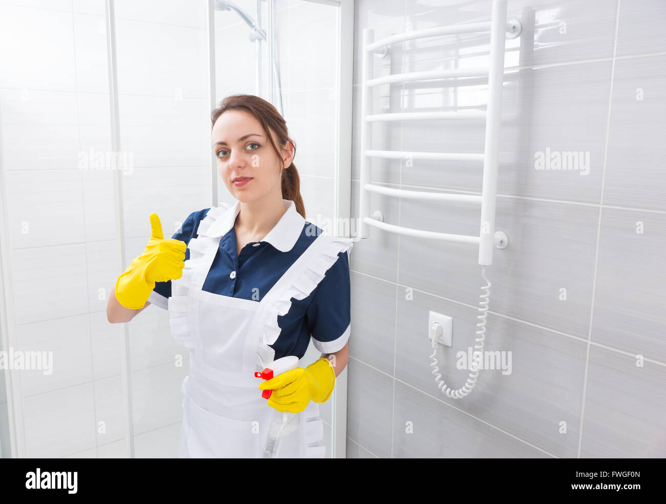 Fiducioso femmina e giovane lavoratore domestico in piedi in vestito blu e grembiule bianco con il pollice in alto gesto tenendo la bottiglia spray vicino al box doccia in bagno. Foto Stock