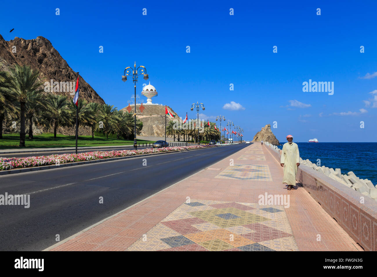 Uomo che indossa dishdasha passeggiate lungo la Mutrah Corniche con bandiere nazionali, letti di fiori e incenso gigante bruciatore, Muscat Oman Foto Stock