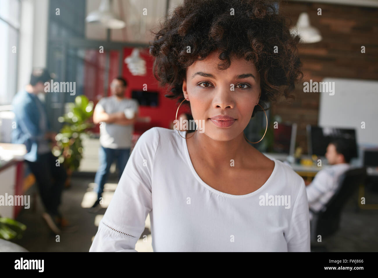 Ritratto di un fiducioso giovani designer femmina in piedi nel suo ufficio con alcuni colleghi in background. Giovane donna africana Foto Stock