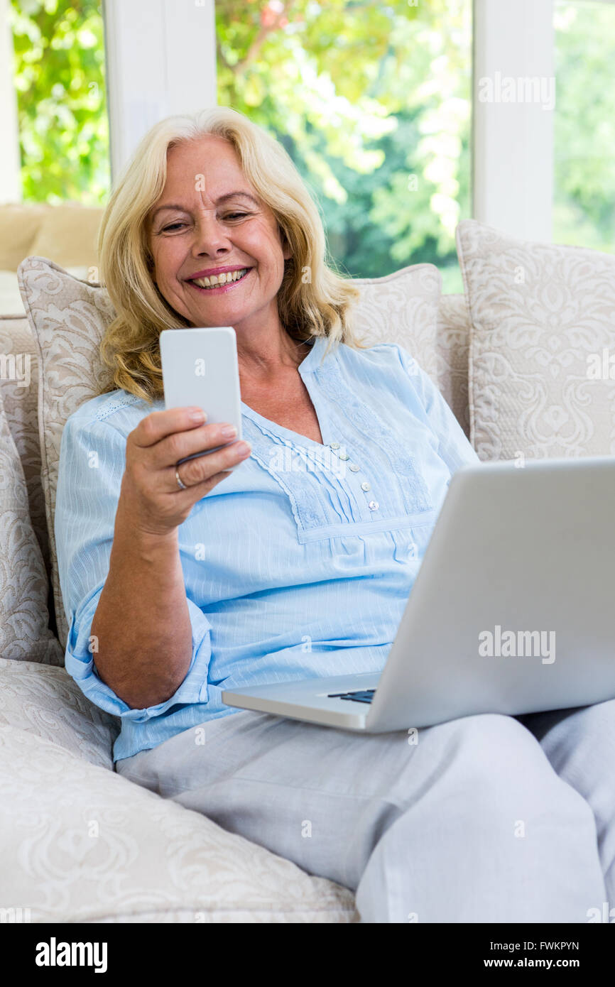 Senior donna utilizzando il telefono cellulare mentre si lavora sul portatile a casa Foto Stock