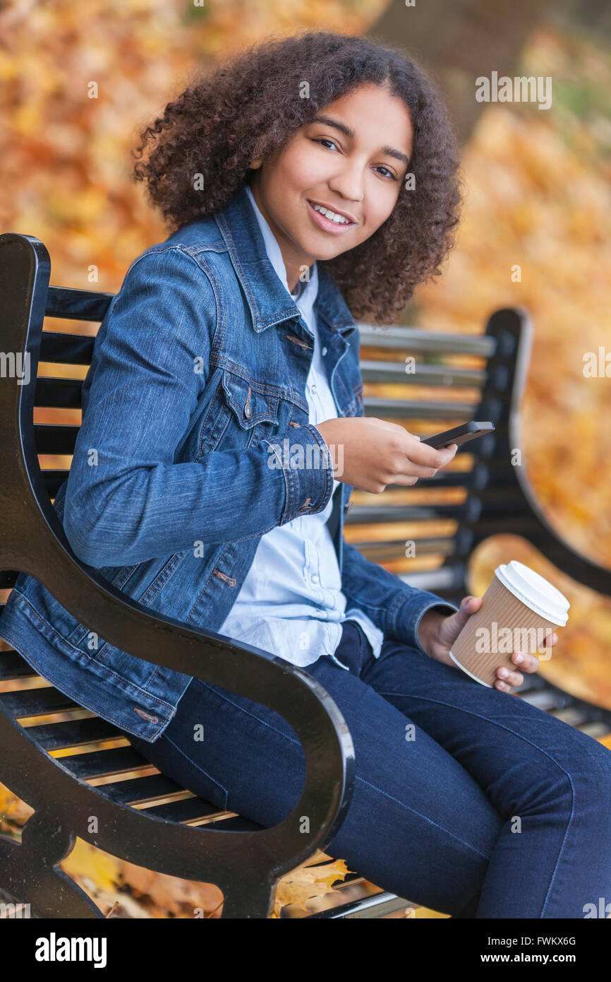 Felice razza mista americano africano ragazza adolescente femmina giovane donna bere take away caffè & sms sul cellulare al di fuori Foto Stock