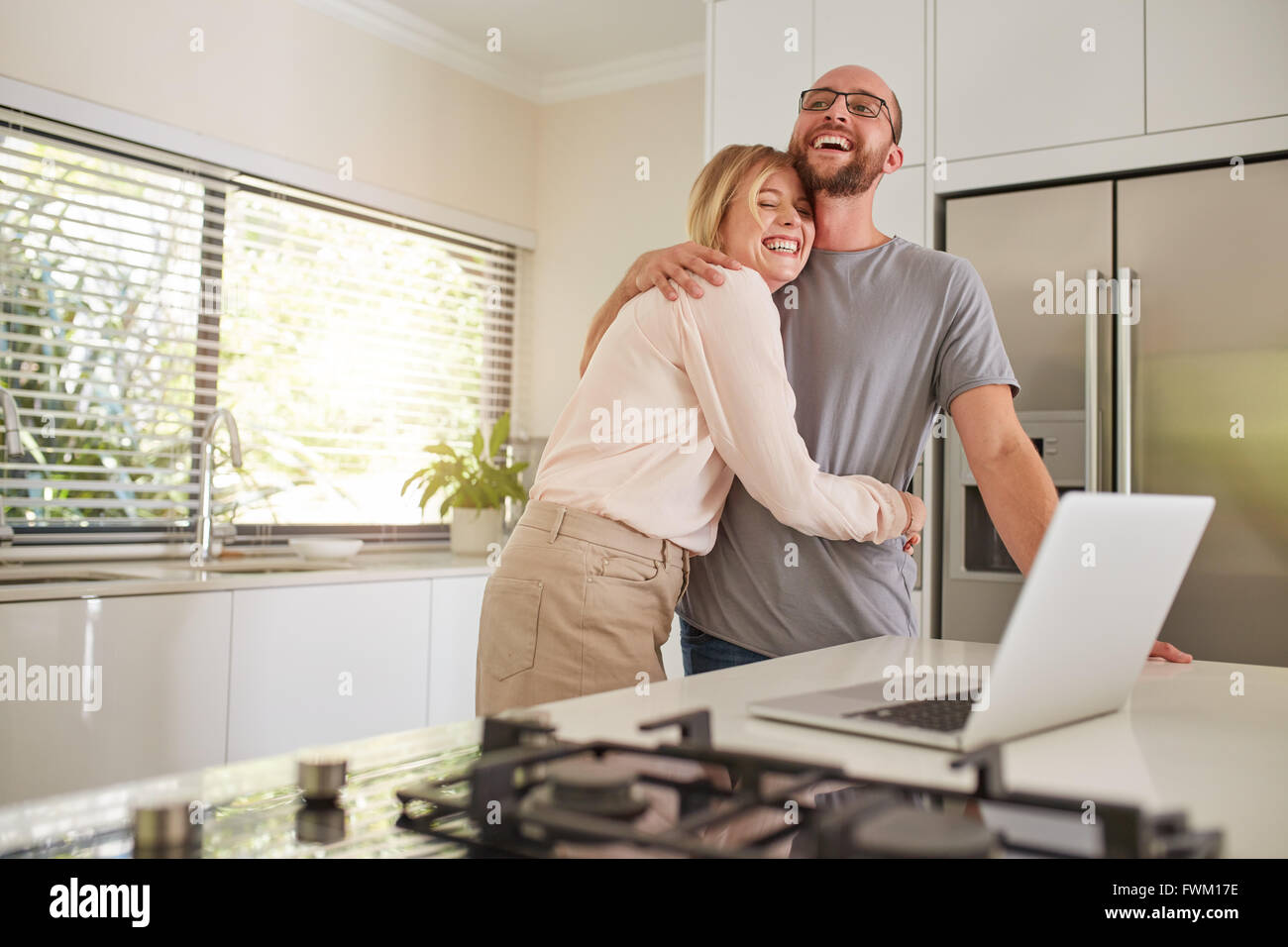 Ritratto di donna felice abbracciando il marito in cucina. Amare giovane con computer portatile sul banco di cucina a casa. Foto Stock