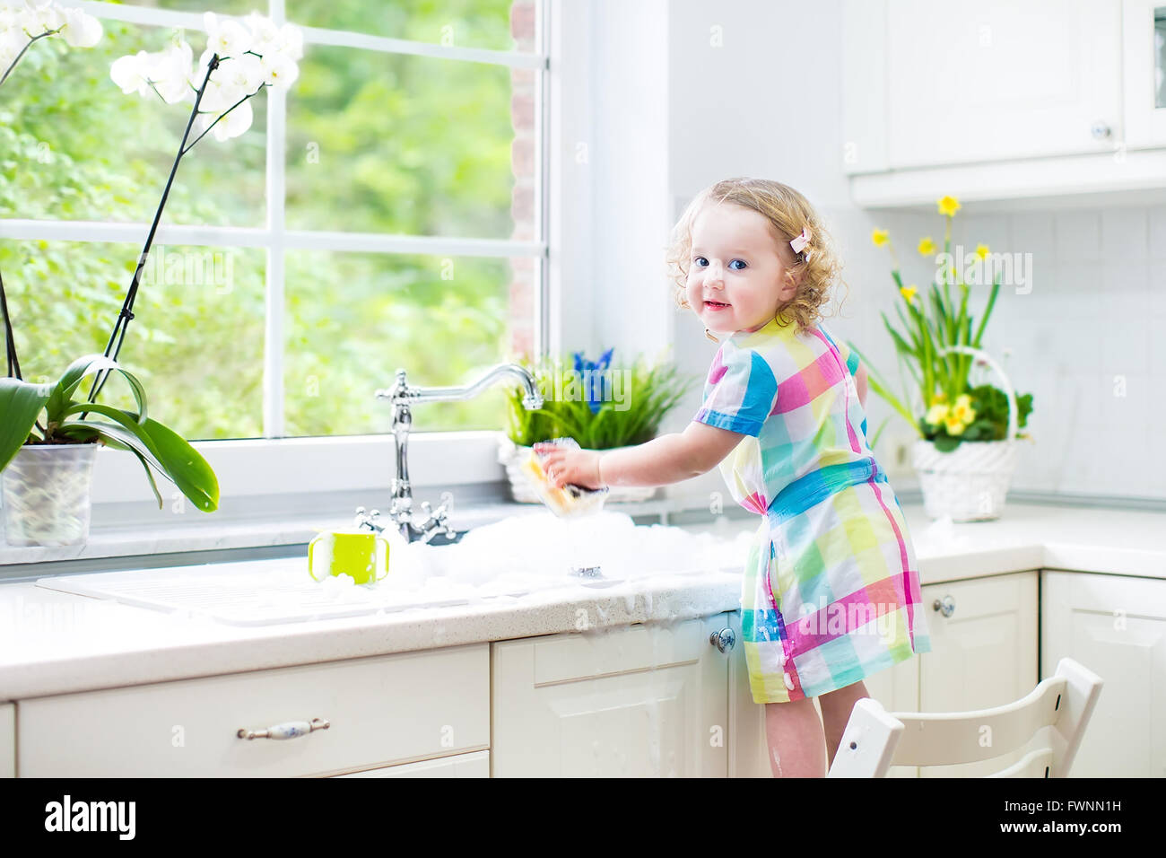 Carino curly toddler ragazza in un abito colorato lavaggio piatti, la pulizia con una spugna e giocare con la schiuma nel lavandino Foto Stock