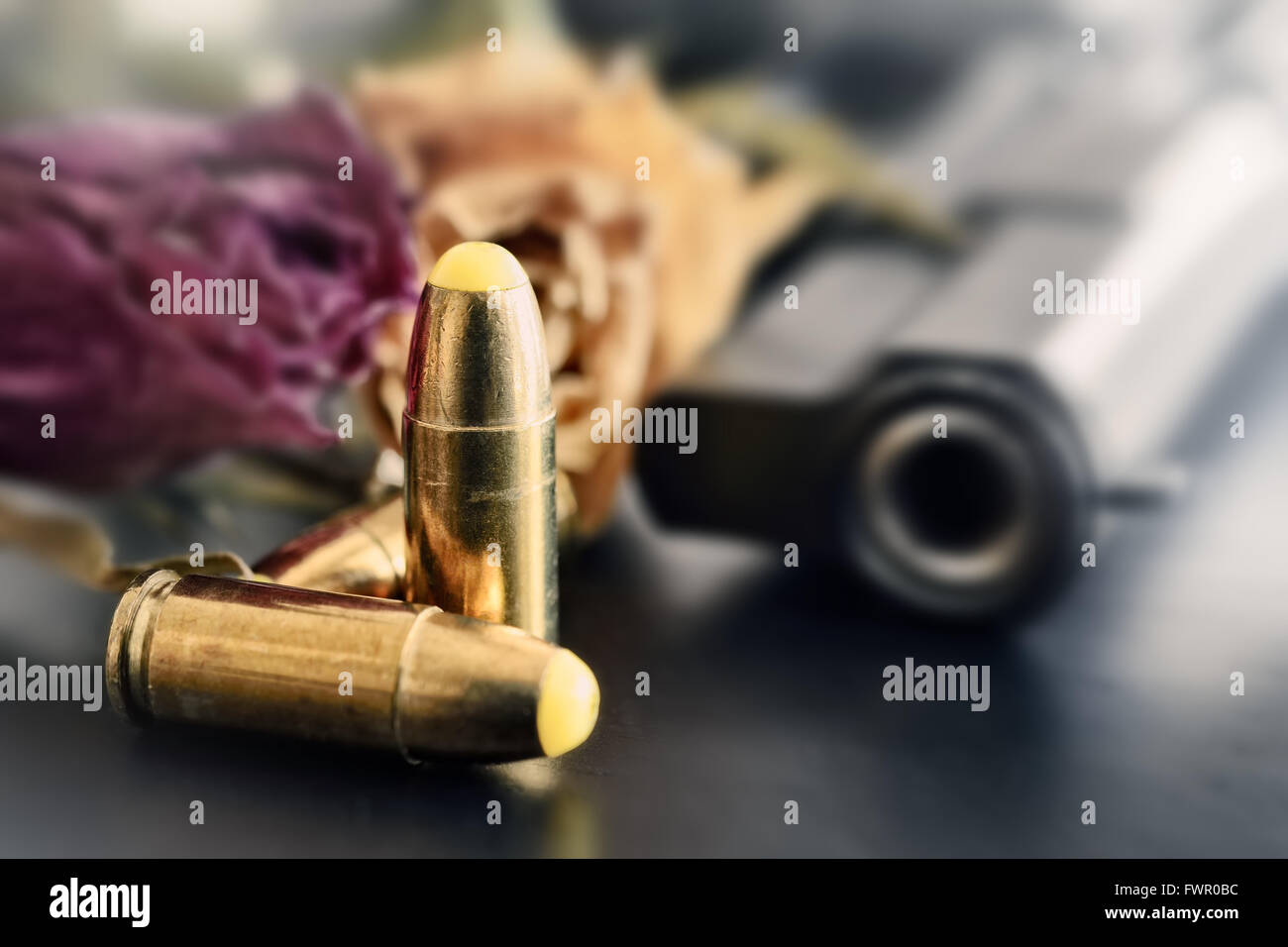 Pistola, proiettili e morti rose fiori su un tavolo scuro contro il sole Foto Stock