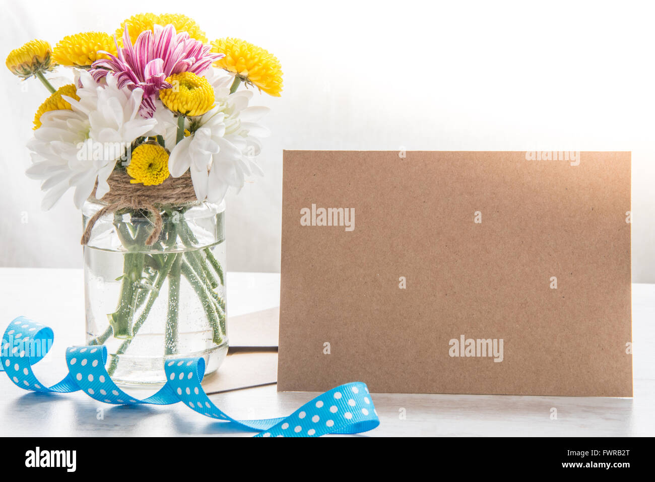 La molla bouquet di fiori in un vaso vaso con carta kraft nota, Busta e puntini blu nastro su un legno bianco sfondo rustico Foto Stock