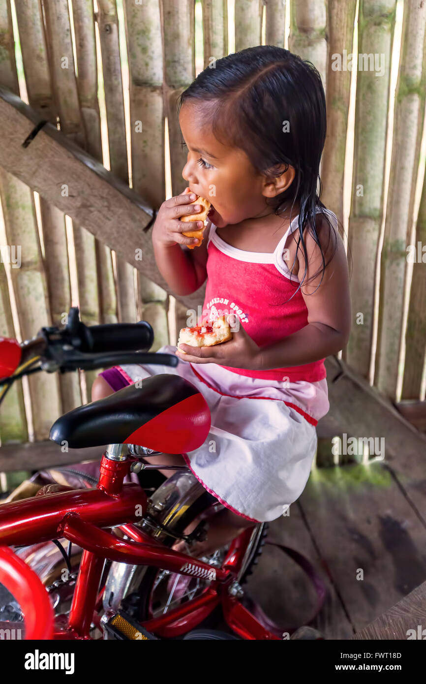 Cuyabeno, Ecuador - 20 Marzo 2015: Ragazza indigena Stand su una bici e mangiare un pezzo di pane con la marmellata in Cuyabeno su Marzo 20 Foto Stock