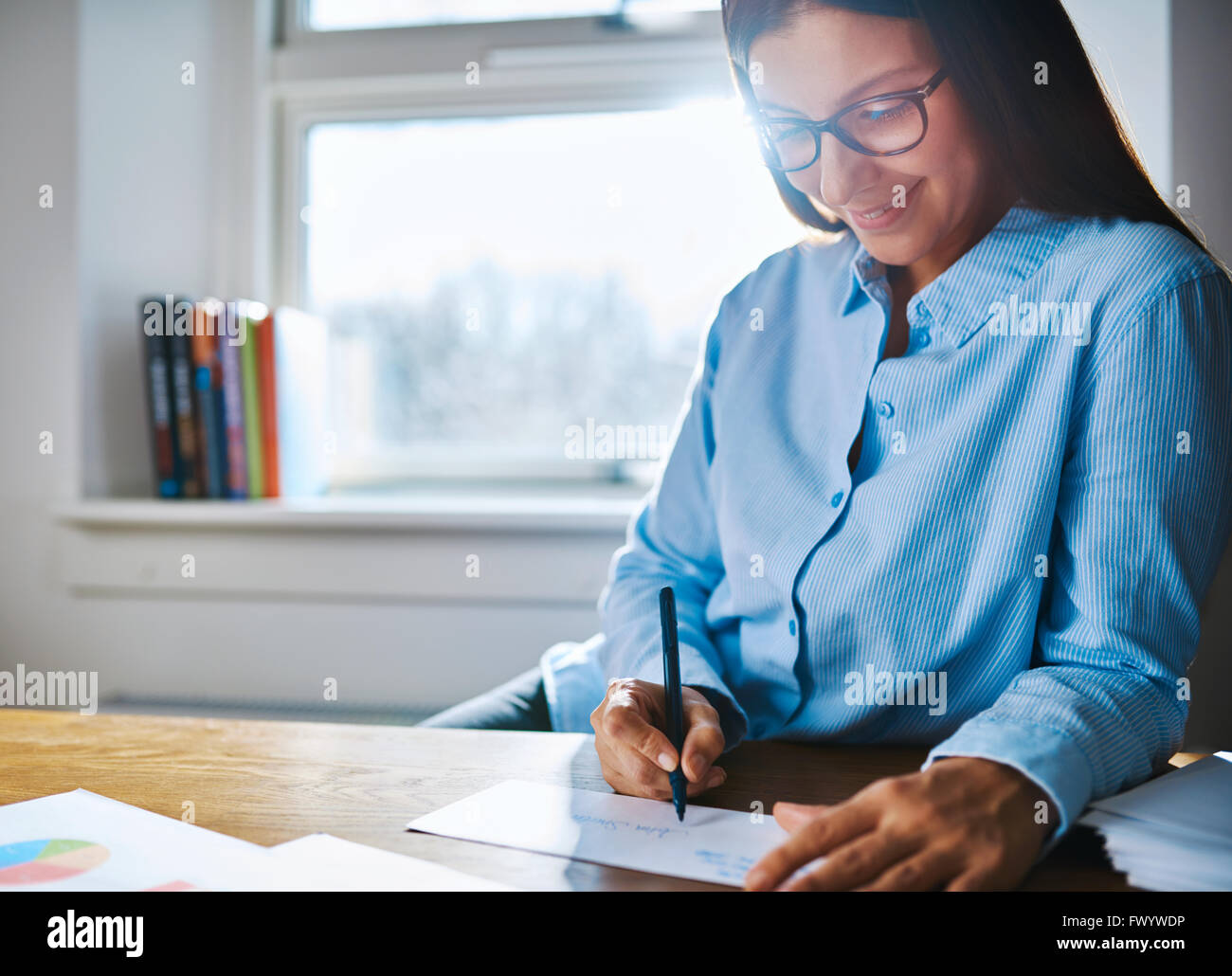 Messa a fuoco selettiva vicino sul felice femmina imprenditore con gli occhiali e la maglietta blu al banco la scrittura sul modulo accanto alla tazza di coff Foto Stock
