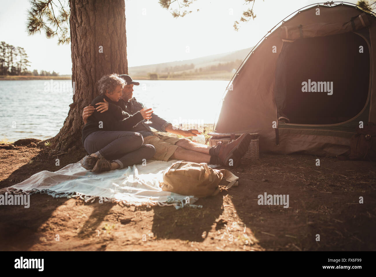 Ritratto di amorevole coppia senior campeggio nei pressi di un lago in una bella giornata d'estate. Essi sono qui seduti sotto un albero al di fuori della tenda, Foto Stock