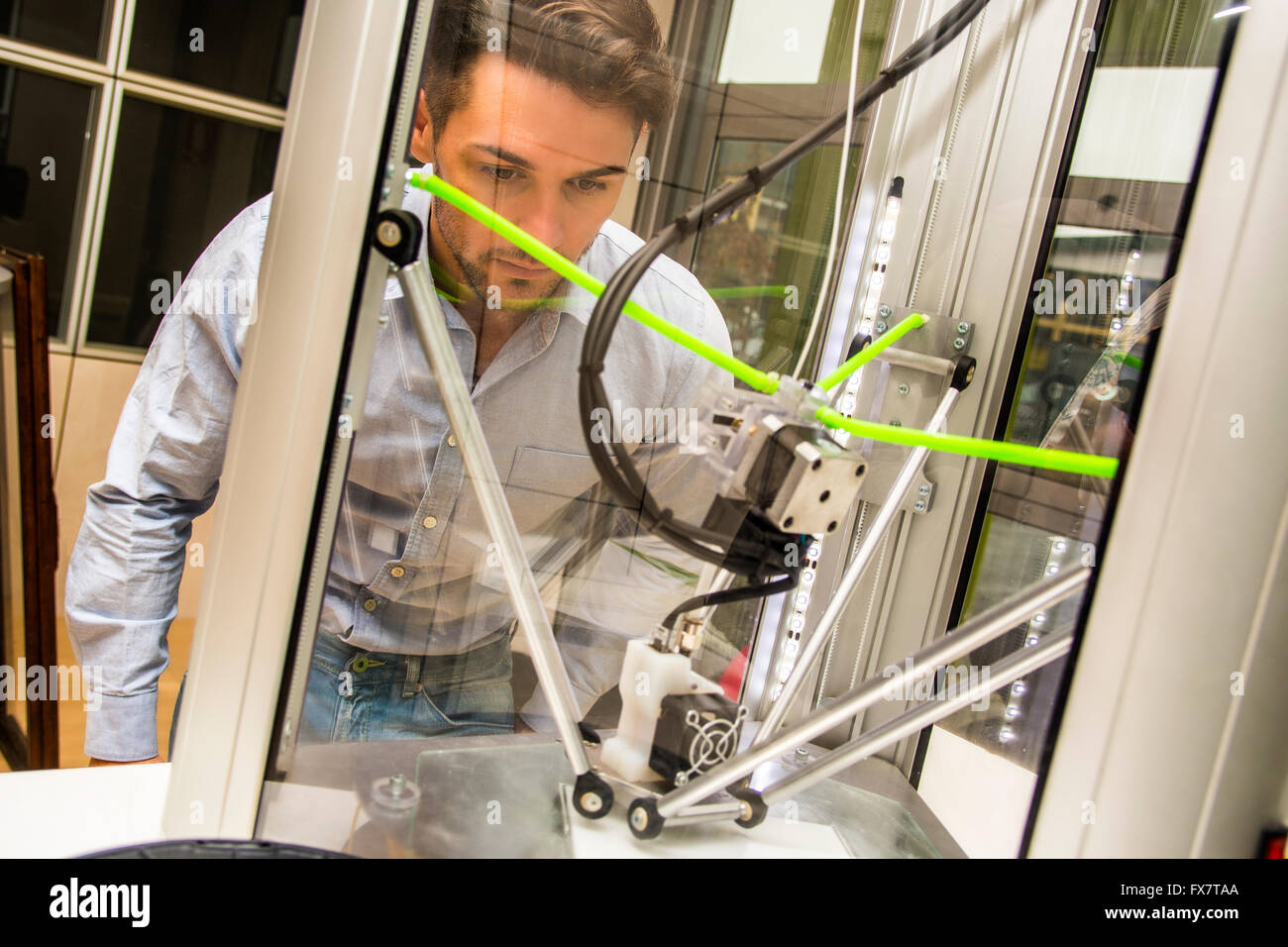 Brunet adulto uomo lavora con stampante 3D, osservando la macchina sul luogo di lavoro Foto Stock