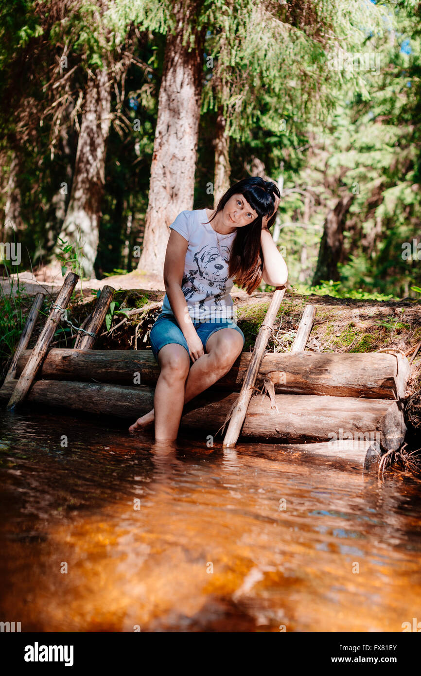 Giovane donna seduta su log e abbassato i suoi piedi nel fiume. Foto Stock
