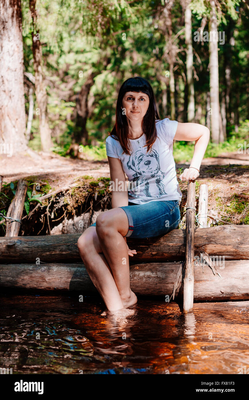 Giovane donna seduta su log e abbassato i suoi piedi nel fiume. Foto Stock