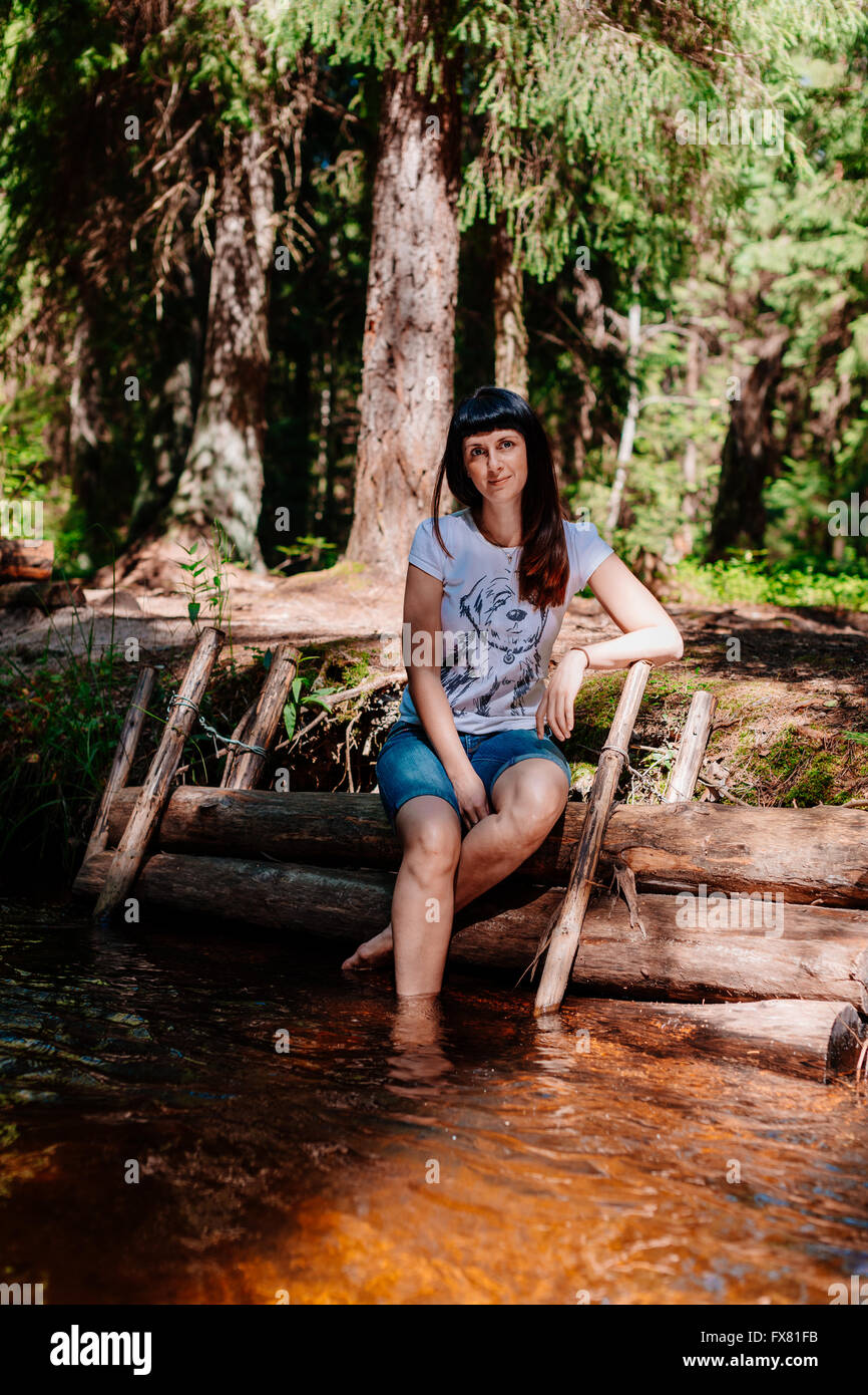 Giovane donna seduta su log e abbassato i suoi piedi nel fiume. Foto Stock