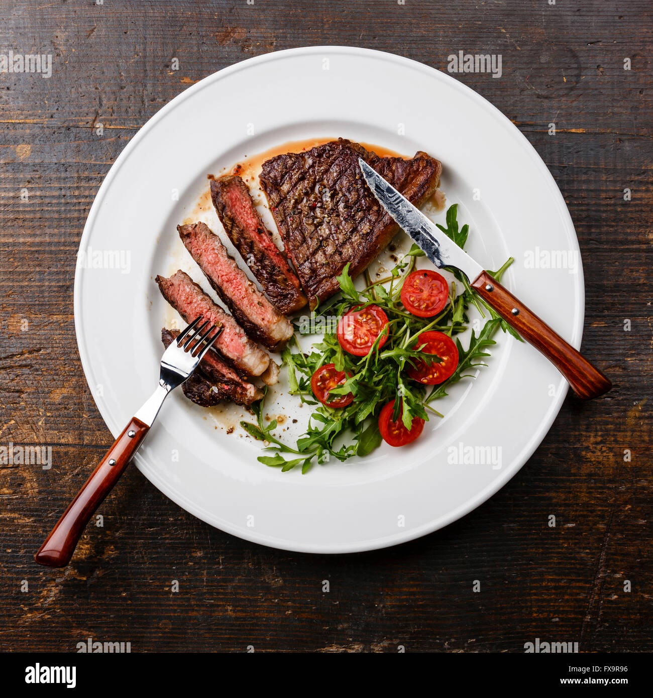 Fette di mezzo raro grigliate di carne di manzo del roastbeef con insalata e pomodori e rucola sulla piastra bianca su scuro dello sfondo in legno Foto Stock