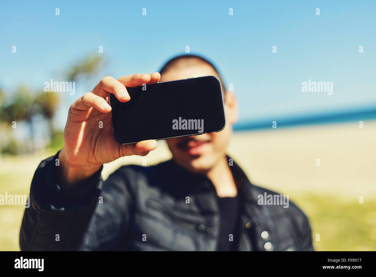 Un giovane uomo che indossa un baffi prende un autoritratto con il suo smartphone di fronte all'oceano Foto Stock