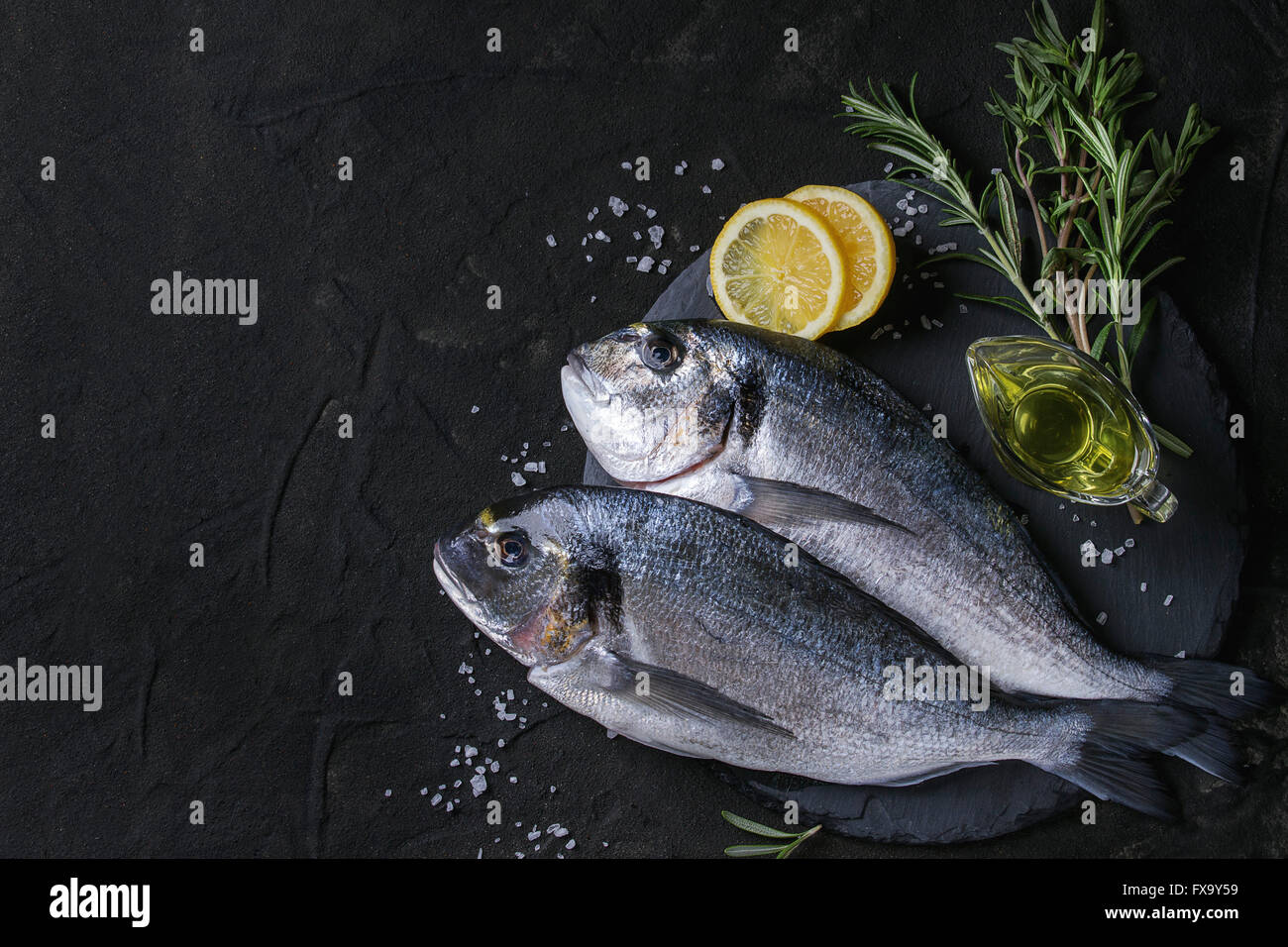 Due pronti per cucinare materie orate di pesce con erbe aromatiche, limone e olio di oliva su pietra ardesia scheda sopra nero testurizzato sfondo. Con copia Foto Stock