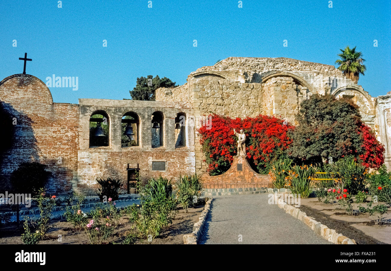 Luminose fiori di bouganville circondano una statua del missionario spagnolo Junipero Serra abbracciando un giovane ragazzo indiano, un monumento del 1914 dall'artista americano Tole Van Rensaalar nel cortile della Missione di San Juan Capistrano in Orange County, California, Stati Uniti d'America. Il leggendario frate francescano fondato la missione nel 1776, settimo del 21 missioni della California ha stabilito in alto e in basso lo stato per espandere della Spagna del territorio e convertire i nativi americani al cristianesimo. Padre Serra è stato canonizzato come santo Cattolica da Papa Francesco nel 2015. Foto Stock