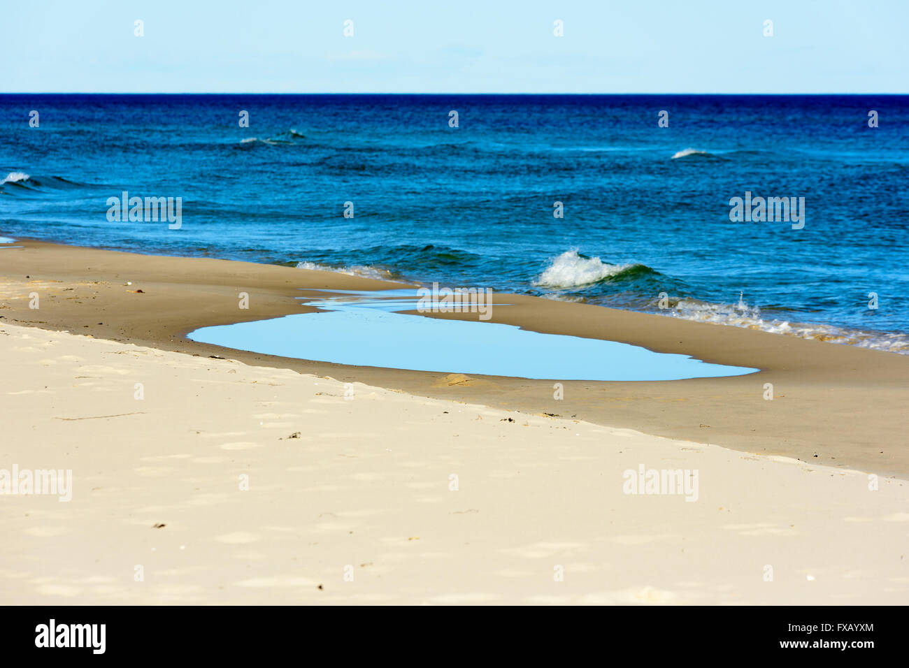 Pozza d'acqua di mare poco profondo sulla spiaggia sabbiosa con il mare aperto in background. Piccole onde alimentare la pozzanghera con nuova acqua. Foto Stock