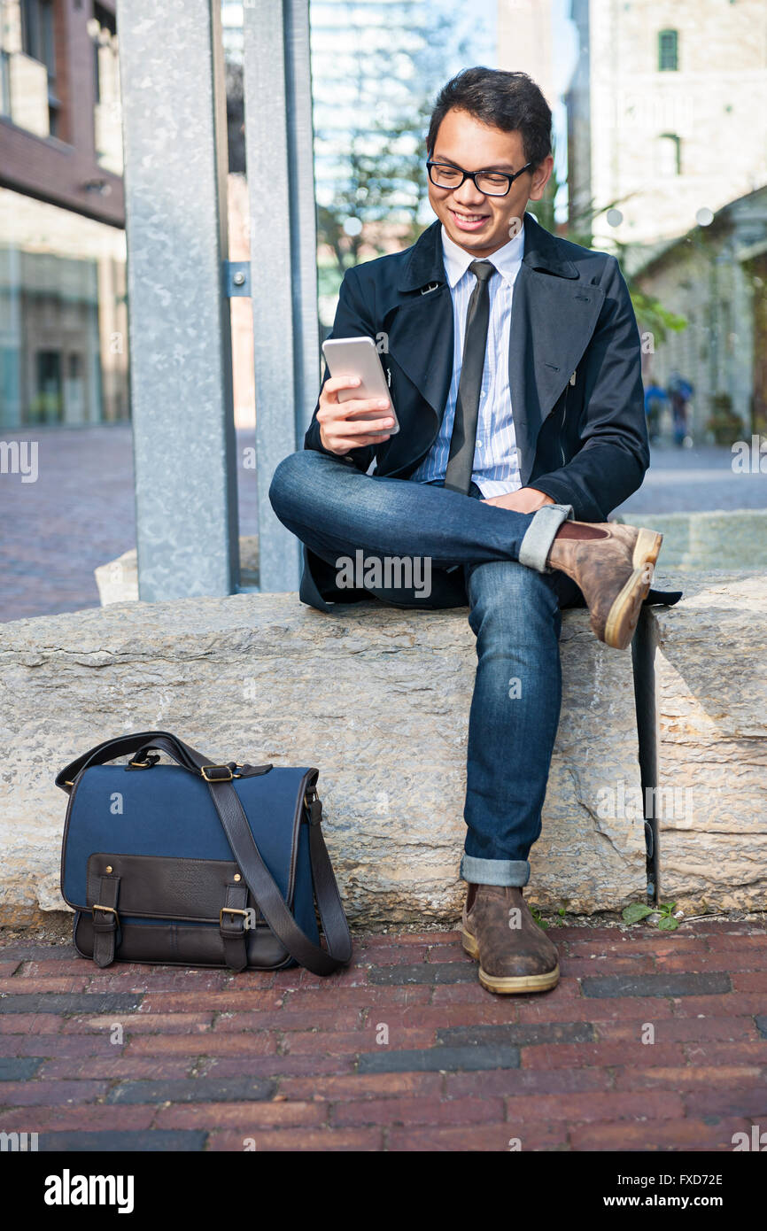 Giovani sorridente asian business un uomo guarda il telefono cellulare al di fuori seduta su una strada di città Foto Stock