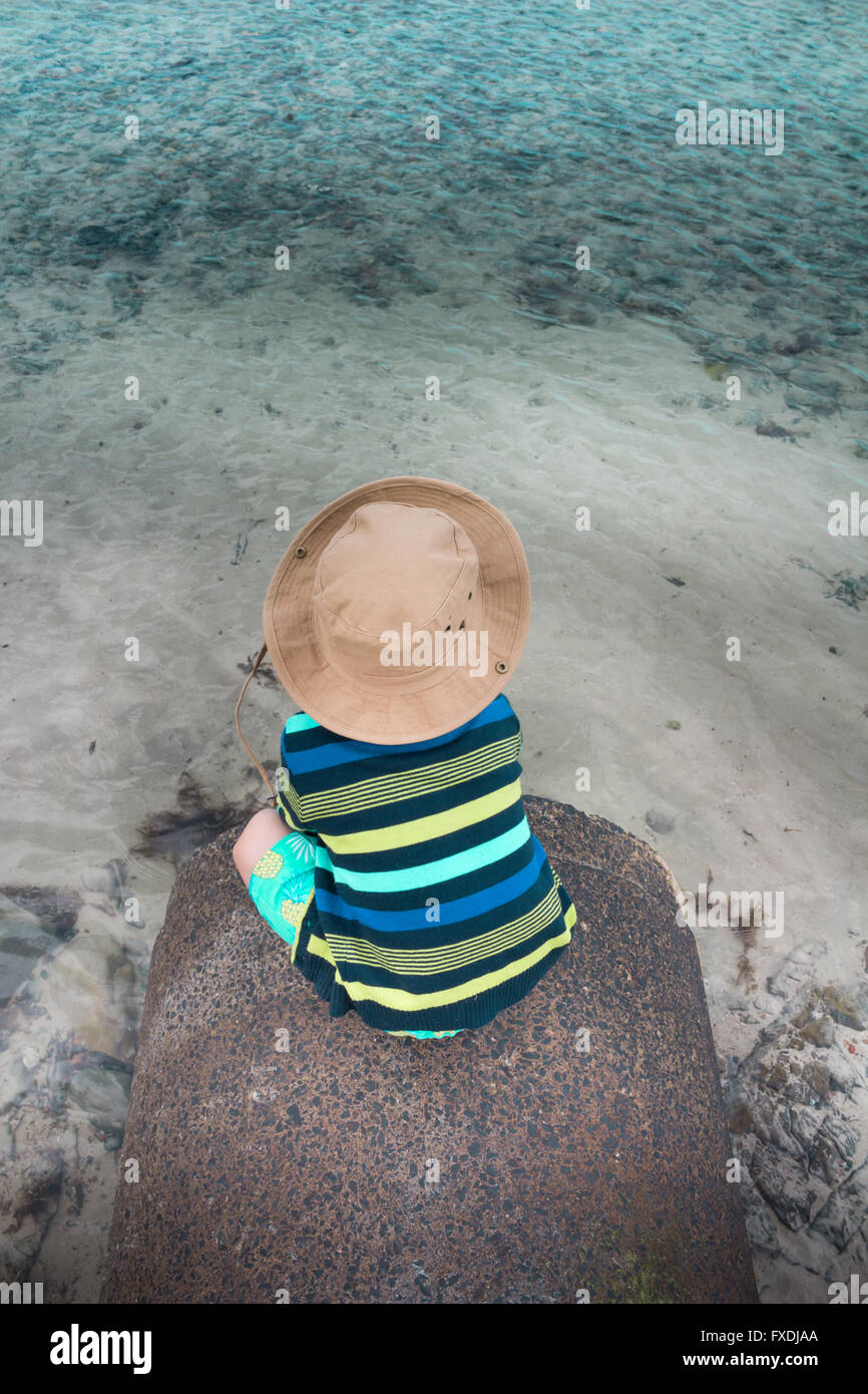 Piccolo Ragazzo seduto sul piccolo molo di pietra con il mare poco profondo e acqua Foto Stock