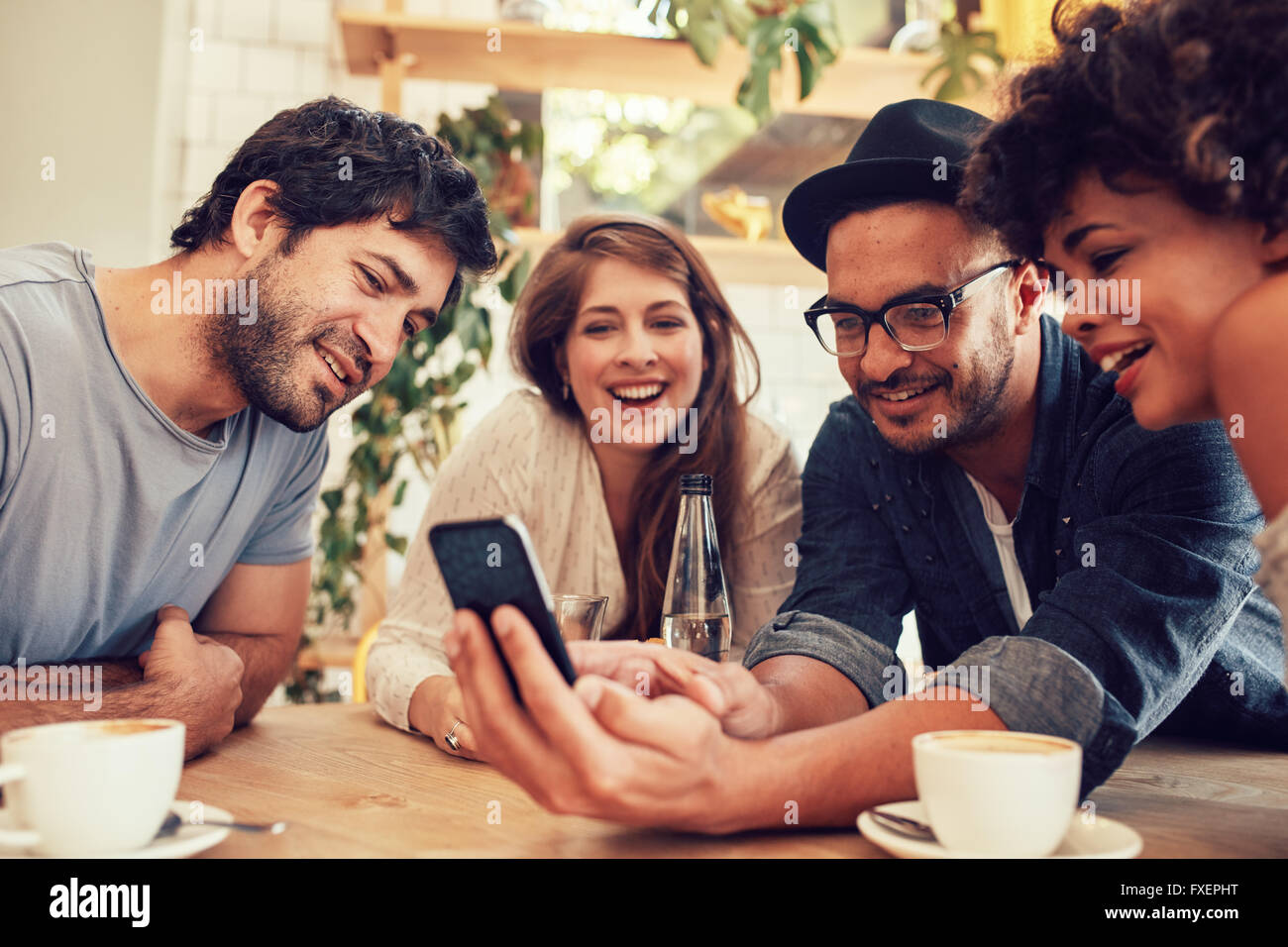 Un gruppo di giovani seduti in un caffè e guardare le foto sul telefono intelligente. Giovani uomini e donne riuniti presso il cafe tabella e Foto Stock