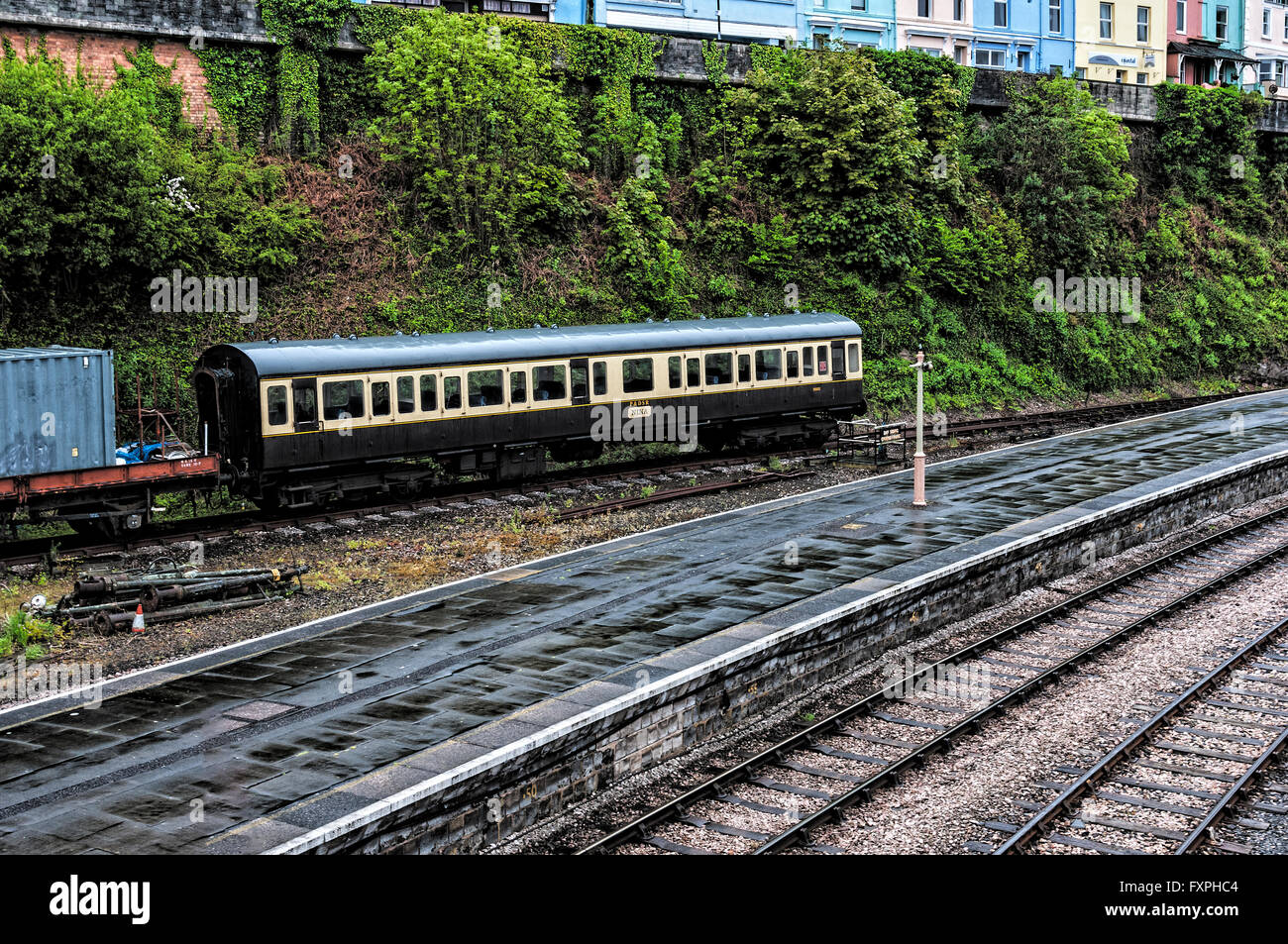 Wet vintage carrozze ferroviarie e di un autocarro a pianale di attendere presso il fondo di un argine dominato da pastello case dipinte Foto Stock