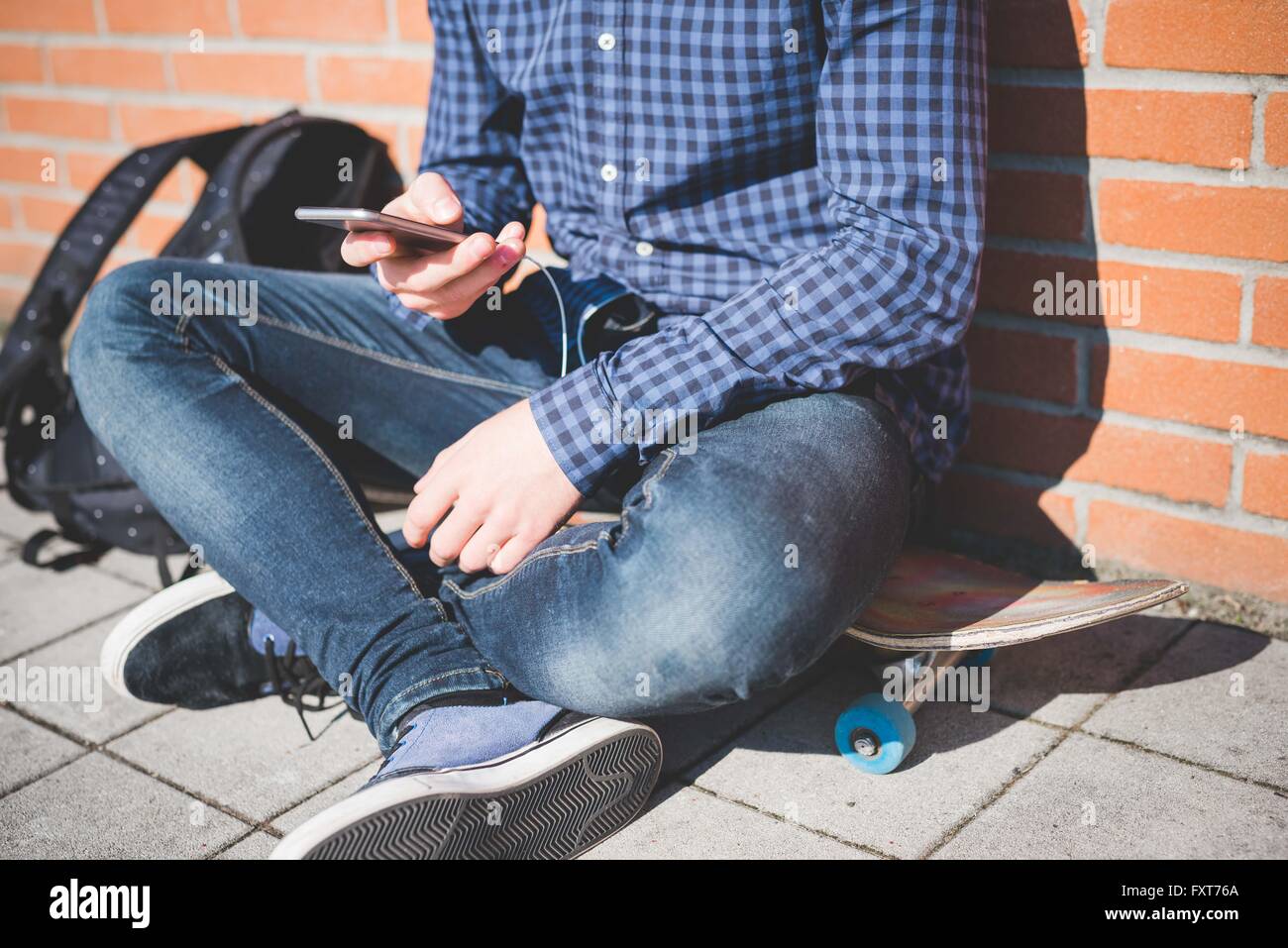 Collo in giù in vista del giovane maschio guidatore di skateboard urbano seduto sul marciapiede di lettura del testo dello smartphone Foto Stock