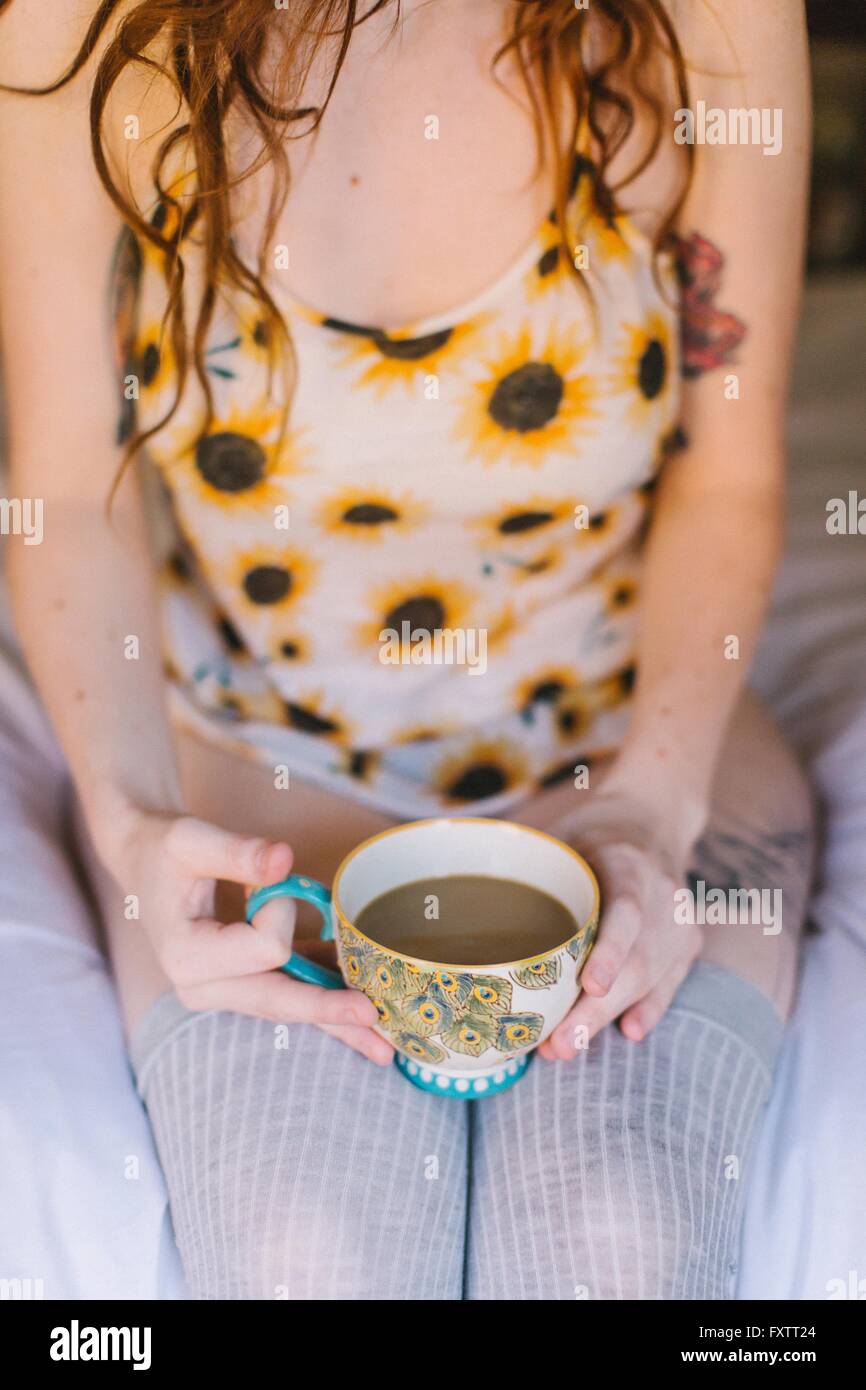 Giovane donna seduta sul letto, tenendo tazzina, metà sezione Foto Stock