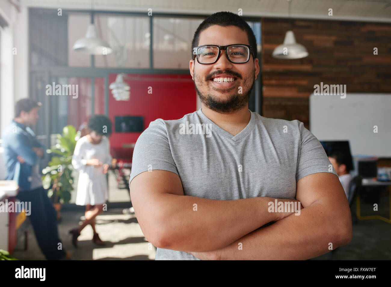 Ritratto di un fiducioso professionista creativo con i colleghi in background. Razza mista uomo in piedi con le braccia incrociate l Foto Stock