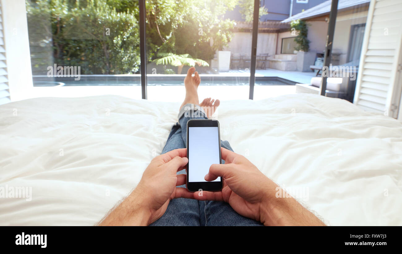 Primo piano immagine della mano umana tenendo il telefono cellulare con schermo vuoto sul letto. POV shot di uomo disteso sul letto tramite smart phone. Foto Stock