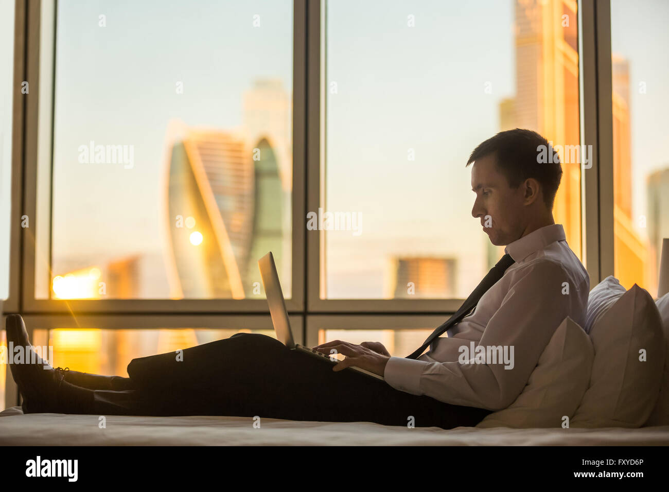 I mediatori che lavorano a casa o in viaggio. Vista laterale verticale del bel giovane imprenditore seduta sul letto e utilizzando computer portatile Foto Stock