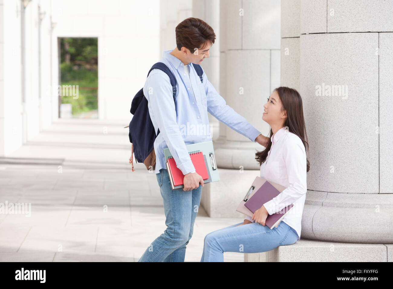 College girl seduti a una colonna dell'edificio del campus e guardando un college boy in piedi accanto a lei con i libri nelle sue mani Foto Stock