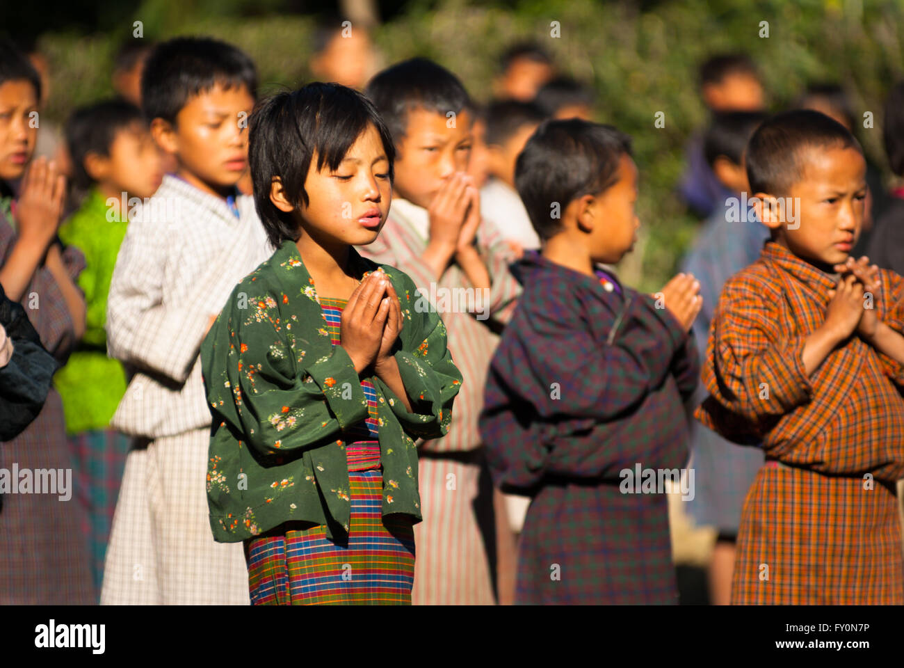 Gli studenti che indossano vestiti tradizionali preghiere al gruppo di mattina in una scuola elementare nel villaggio Nimshong nel sud del Bhutan Foto Stock