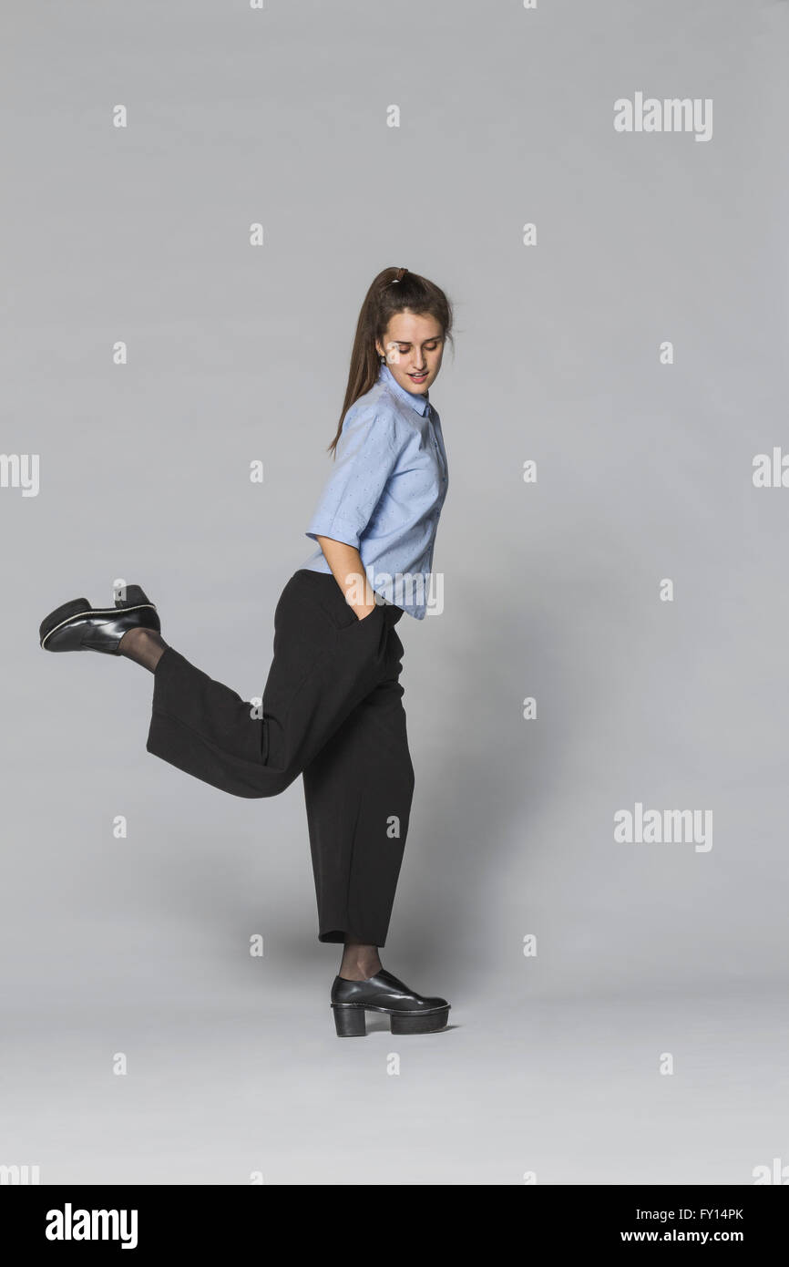 Lunghezza completa donna in piedi su una gamba sola con le mani nelle tasche contro uno sfondo grigio Foto Stock