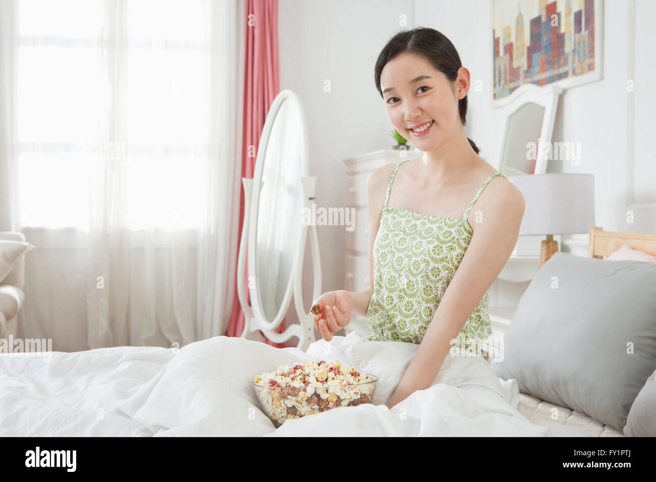 Ritratto di giovane donna sorridente seduta sul letto mangiare popcorn fissando sulla parte anteriore Foto Stock