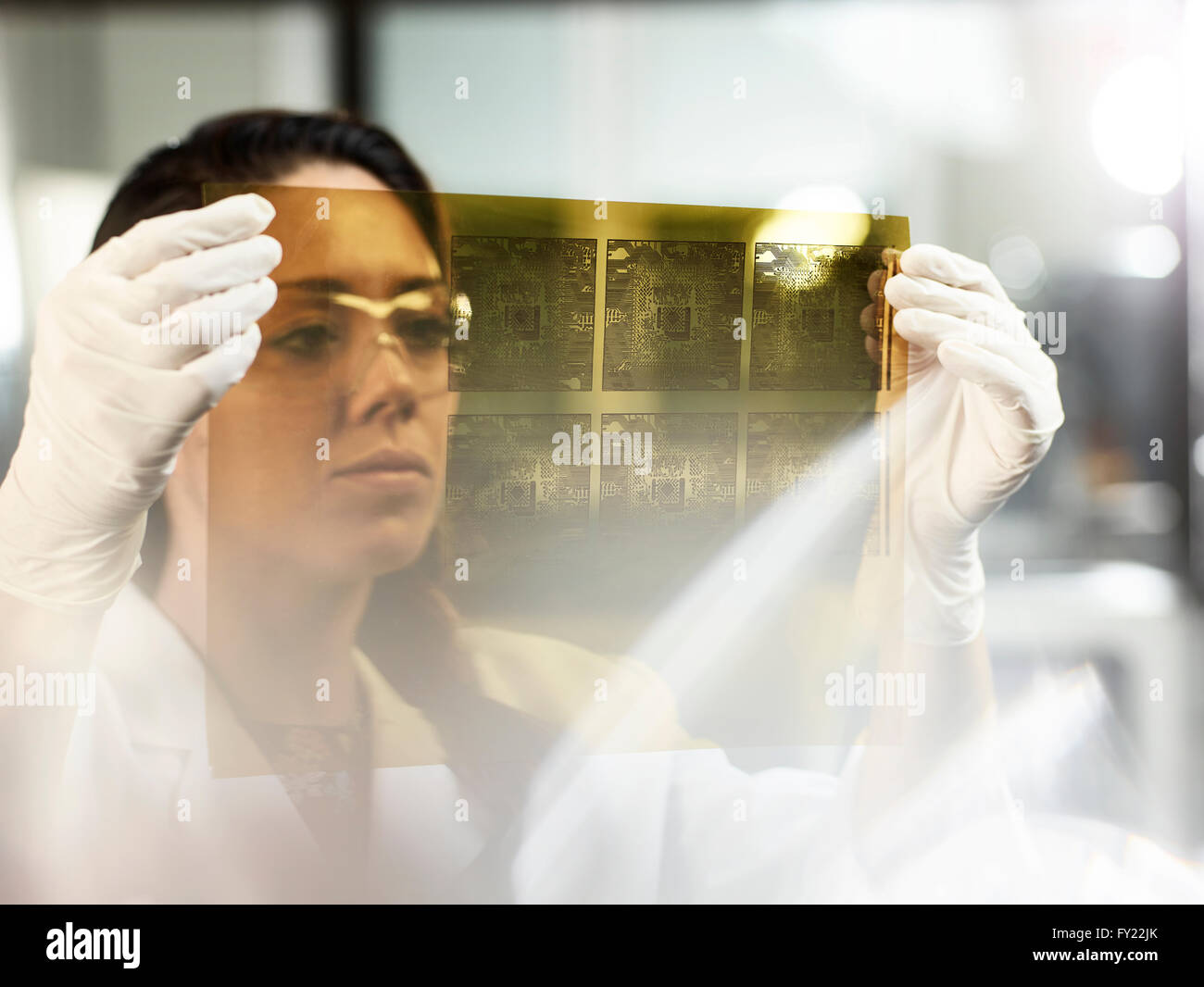 Donna, 25-30 anni, in plastica guanti e occhiali di protezione, controllare i circuiti elettronici stampati su pellicola in un laboratorio di elettronica Foto Stock