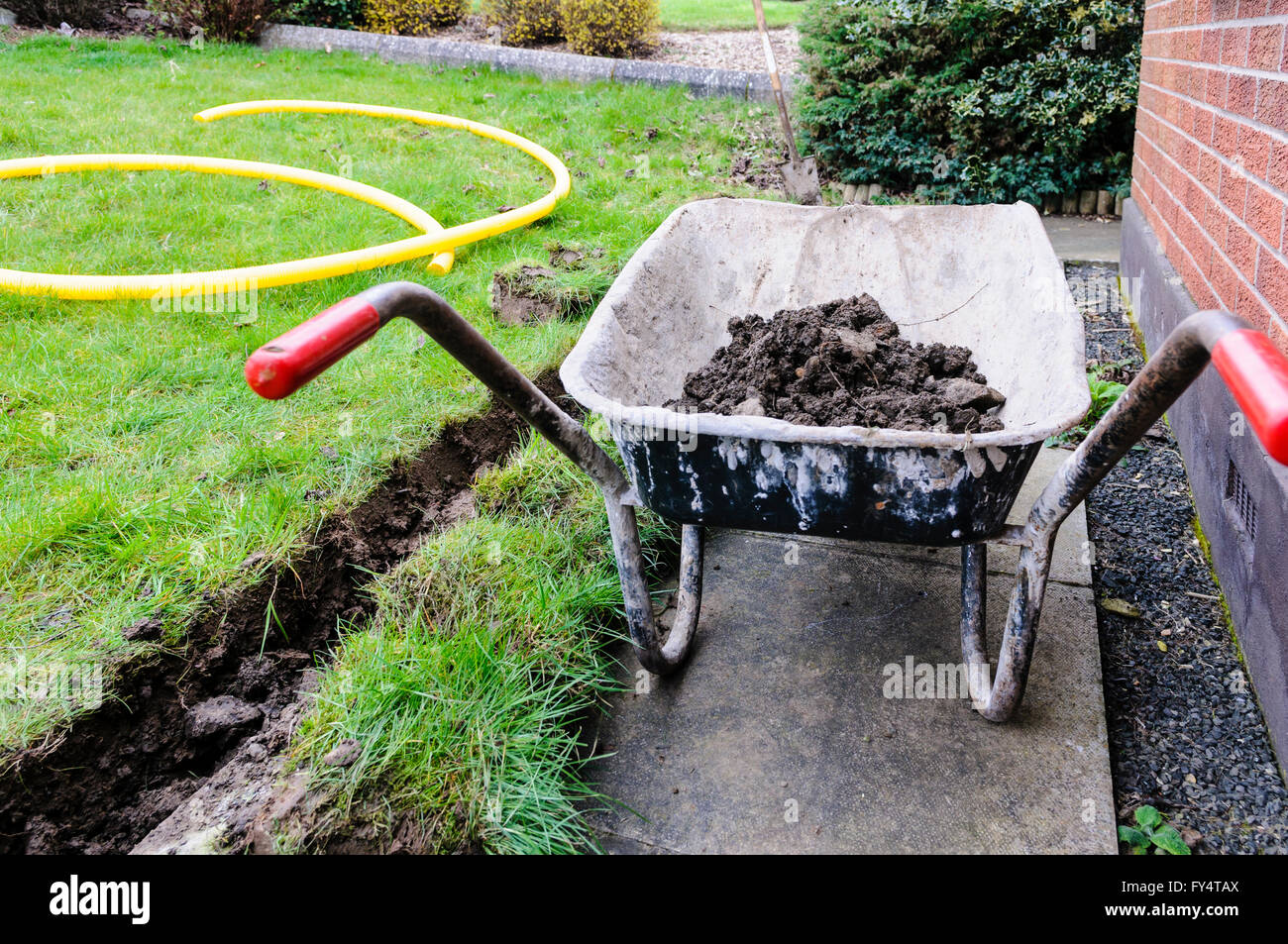 Carriola piena di suolo dopo una trincea è stata scavata in corrispondenza del fondo di un prato per installare un canale di drenaggio. Foto Stock