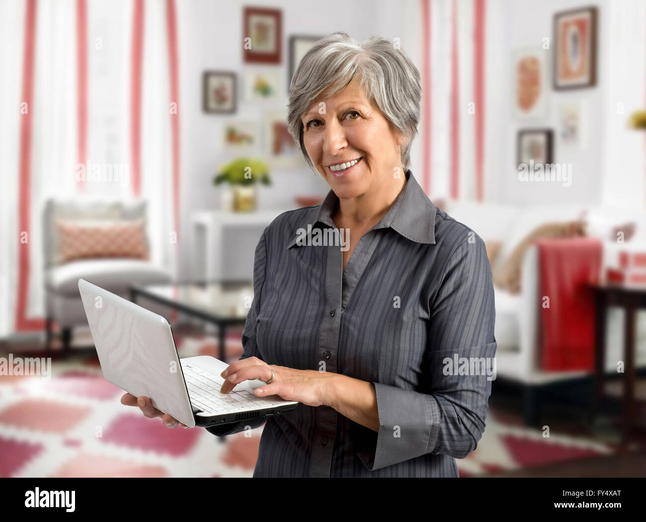 Senior Donna che utilizza il suo computer portatile palmare come essa si erge nel salotto di casa Foto Stock