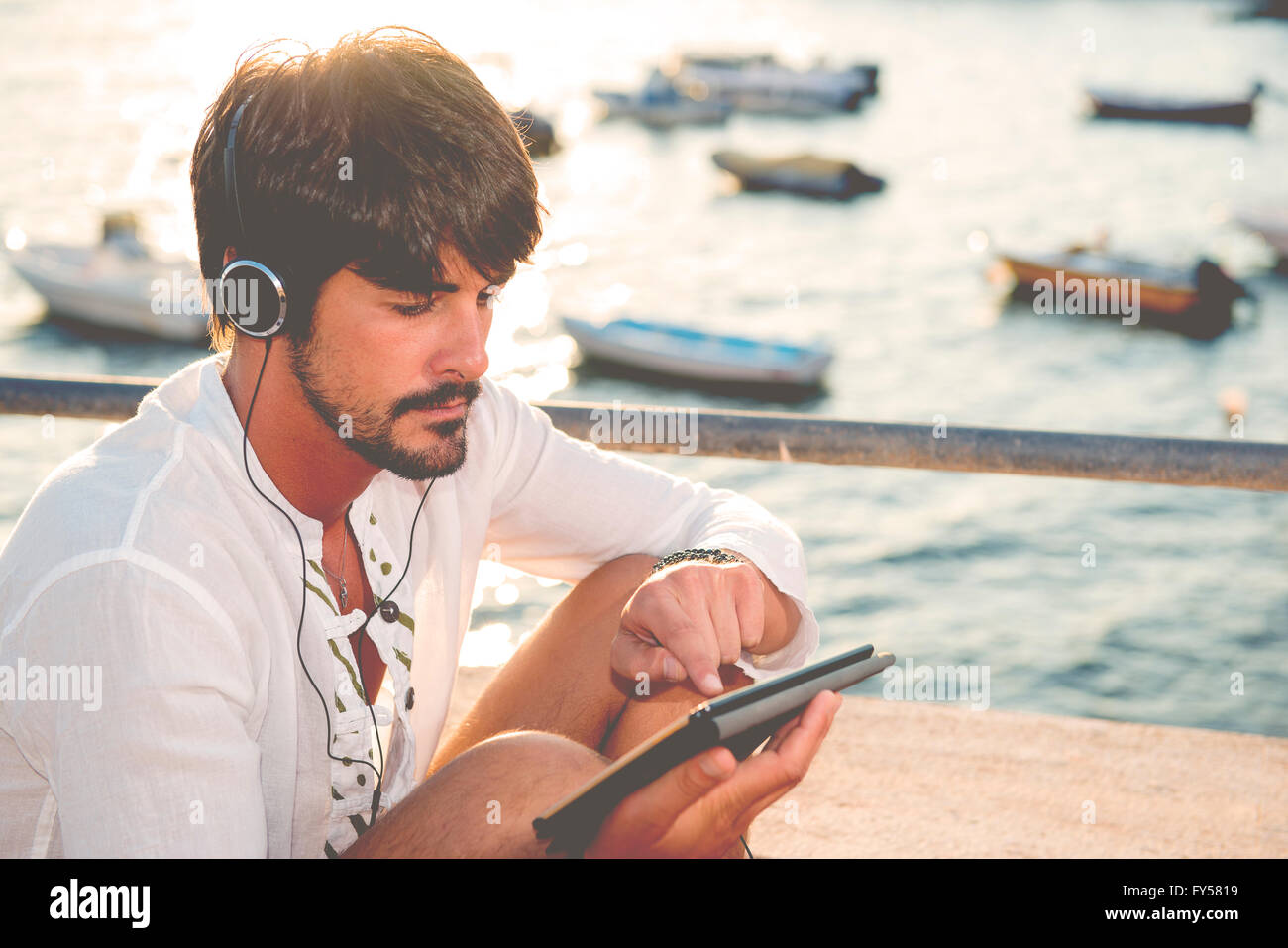 Guy ascoltando musica con le cuffie dalla compressa in una destinazione di vacanze nei pressi di barche Foto Stock