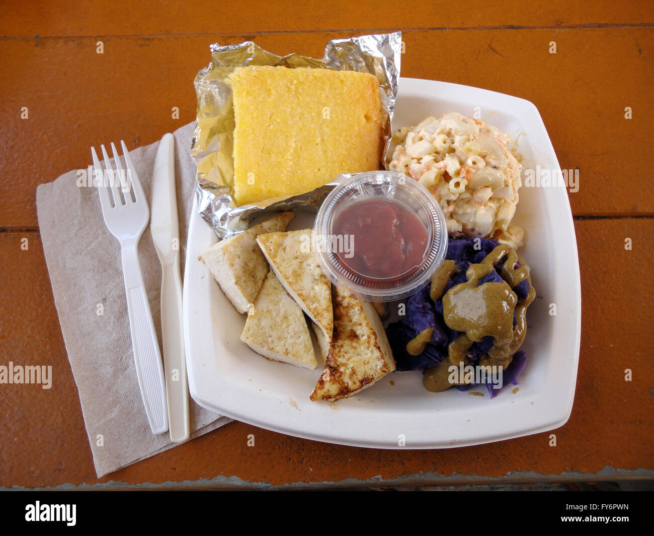 Fette di tofu, patate dolci, il pane di mais e insalata di mac su una piastra quadrata e la forcella di plastica e il coltello su un tavolo da picnic. Foto Stock